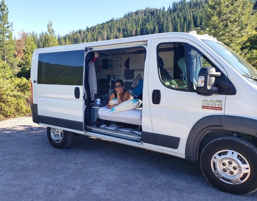 A white cargo van that’s been converted into a camper has its side door open with a woman laying on a bed, looking out the door. The van sits in a forest. 