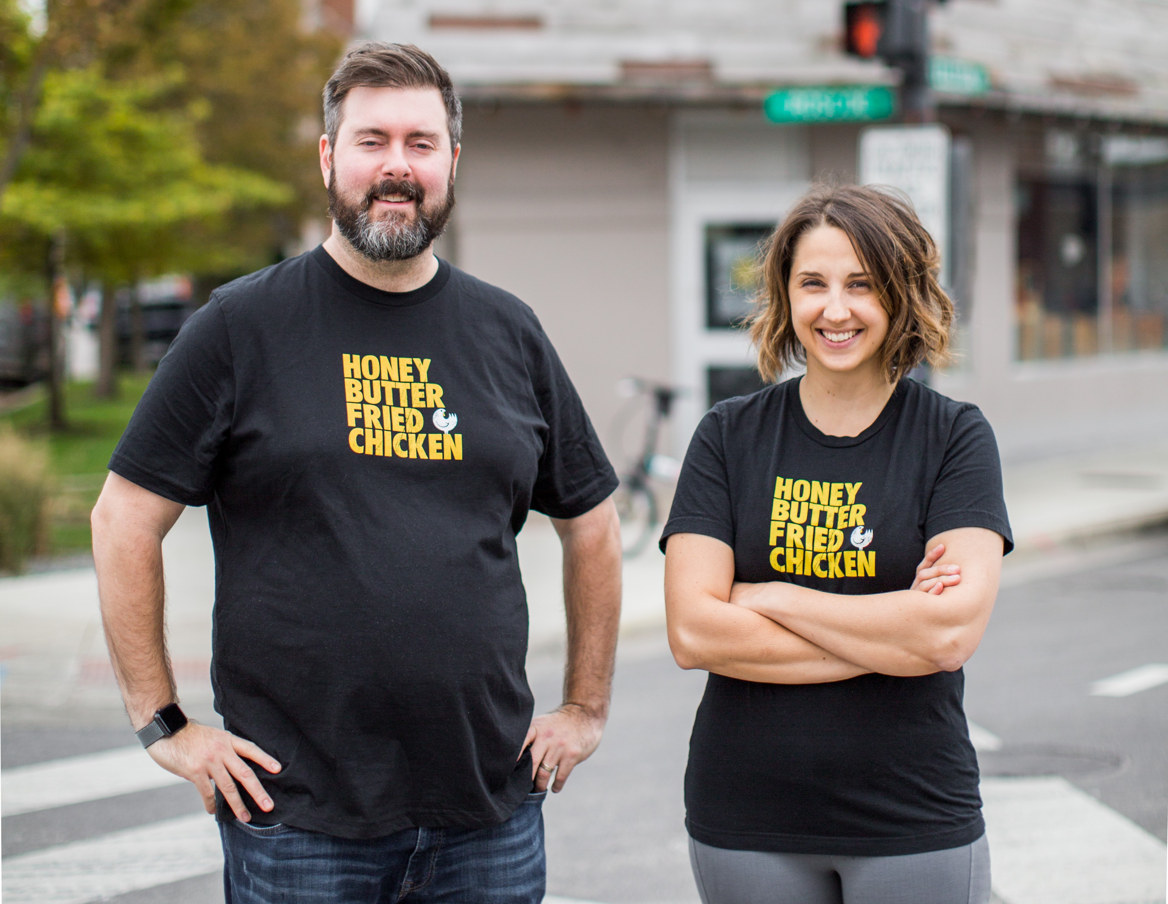 A man and woman wearing the same black T-shirts with the Honey Butter Fried Chicken logo standing outside and smiling.