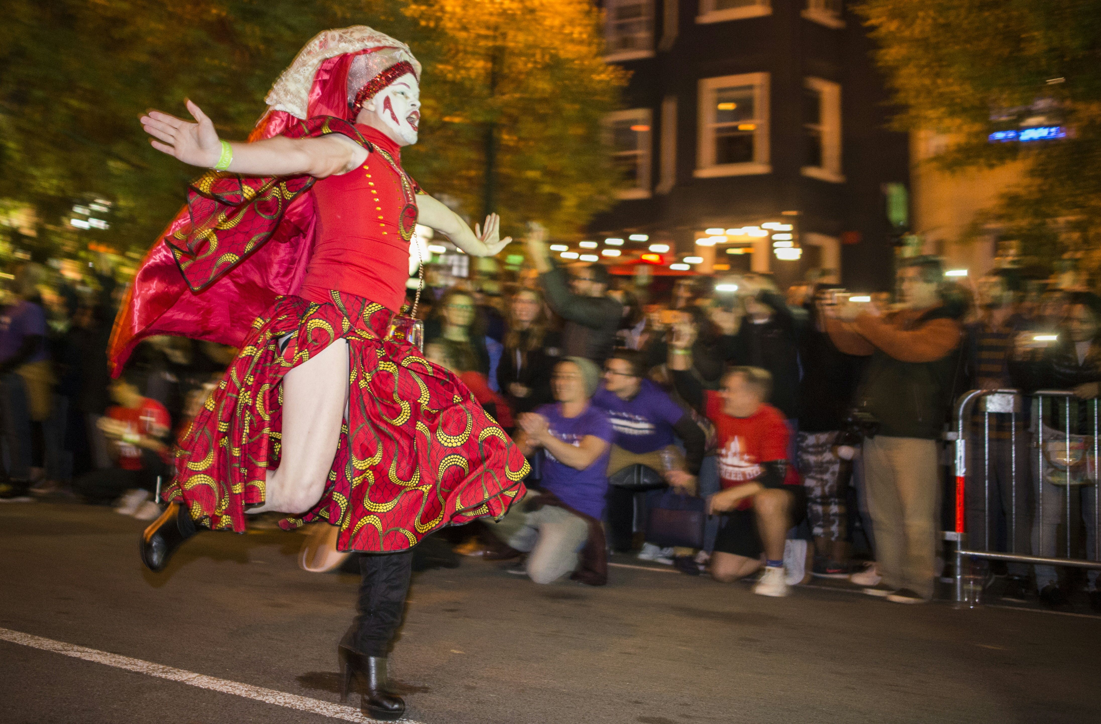 A person running with arms outstretched on a city street. The person is wearing a drag outfit: a red skirt and red top with a red veil. They wear white makeup.