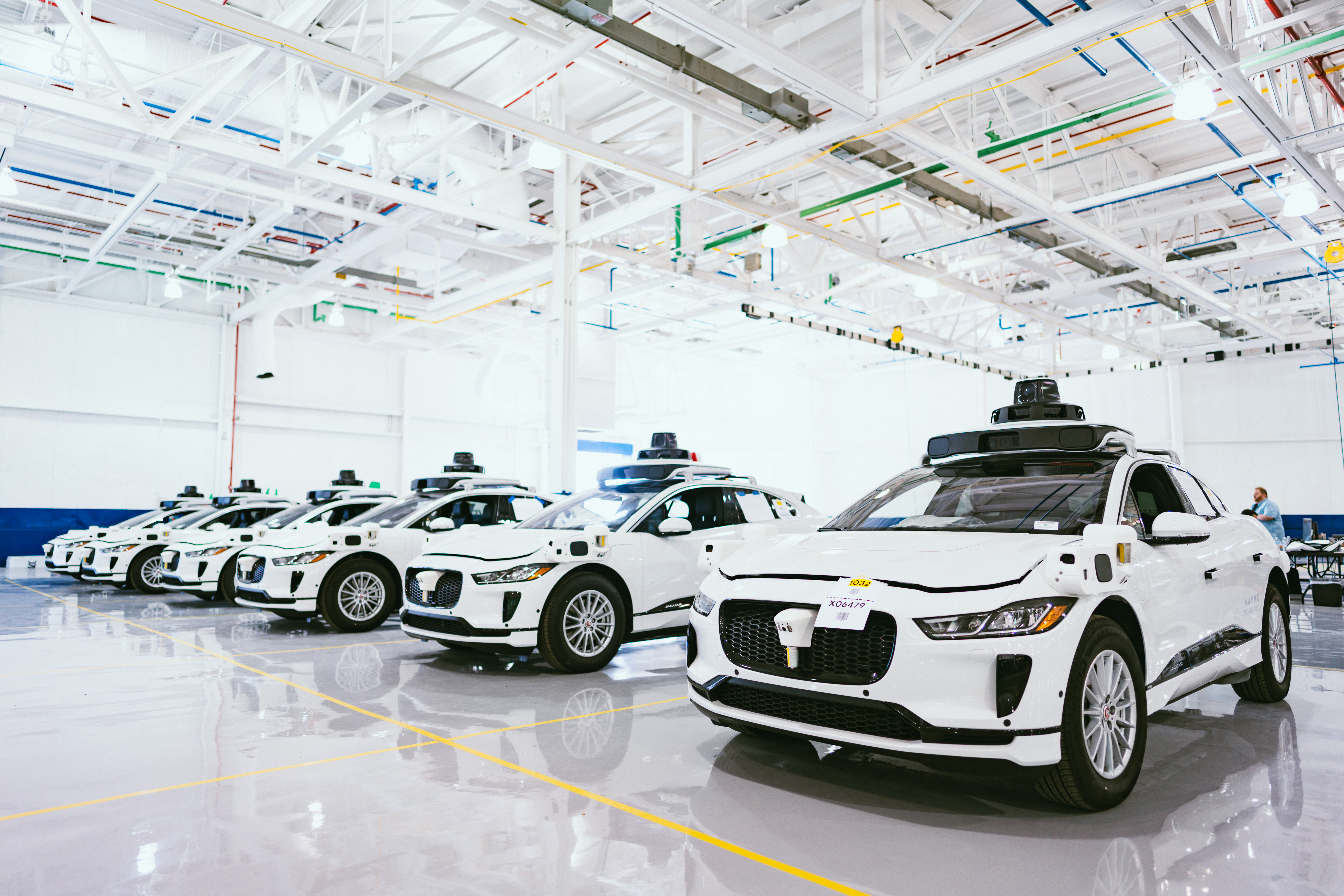 A row of six white sedans idle in a large warehouse with with steel beams in the ceiling. There are black cameras mounted on top of the cars.