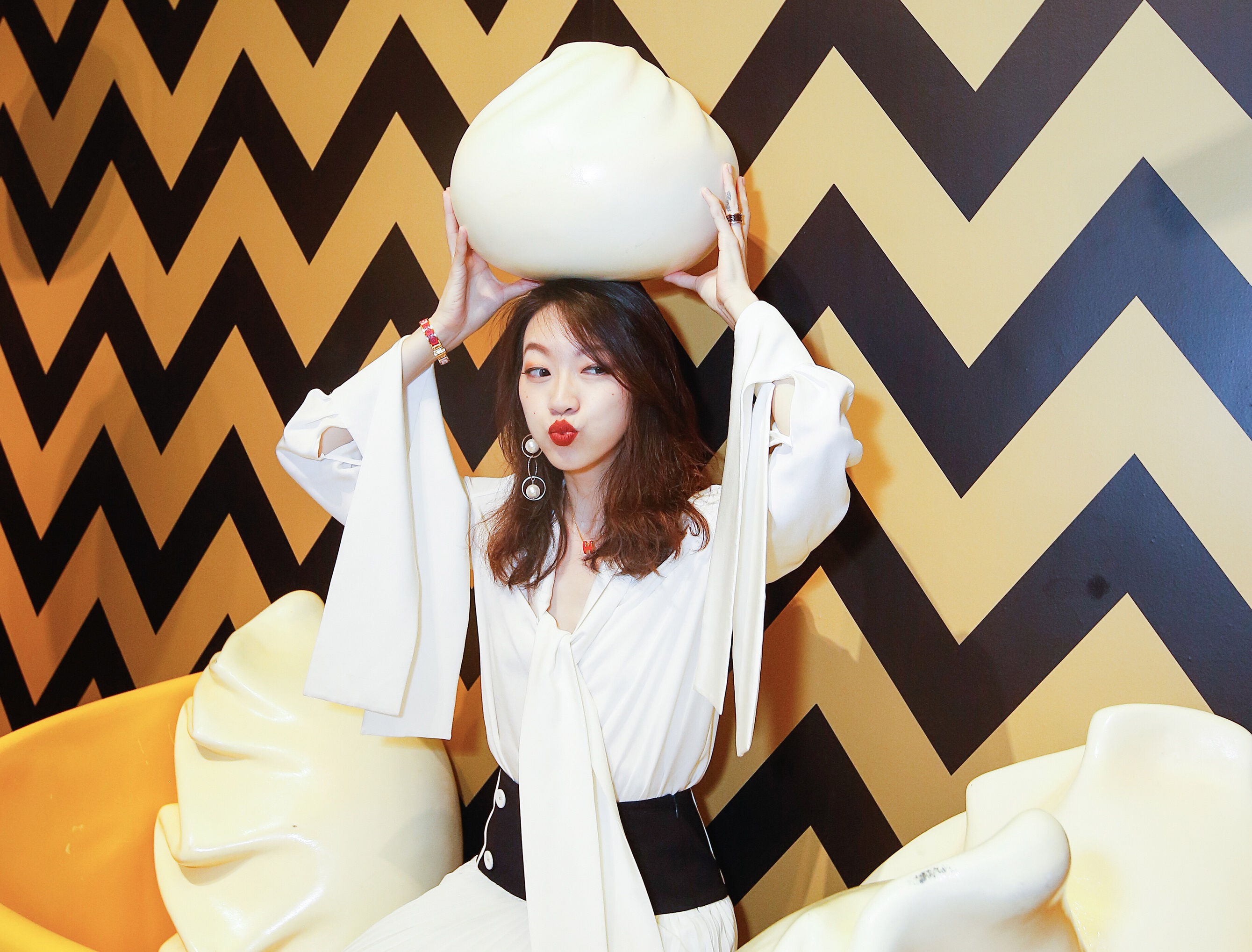 Woman holding a giant fake dumpling in a bowl of dumplings with a black and yellow background.