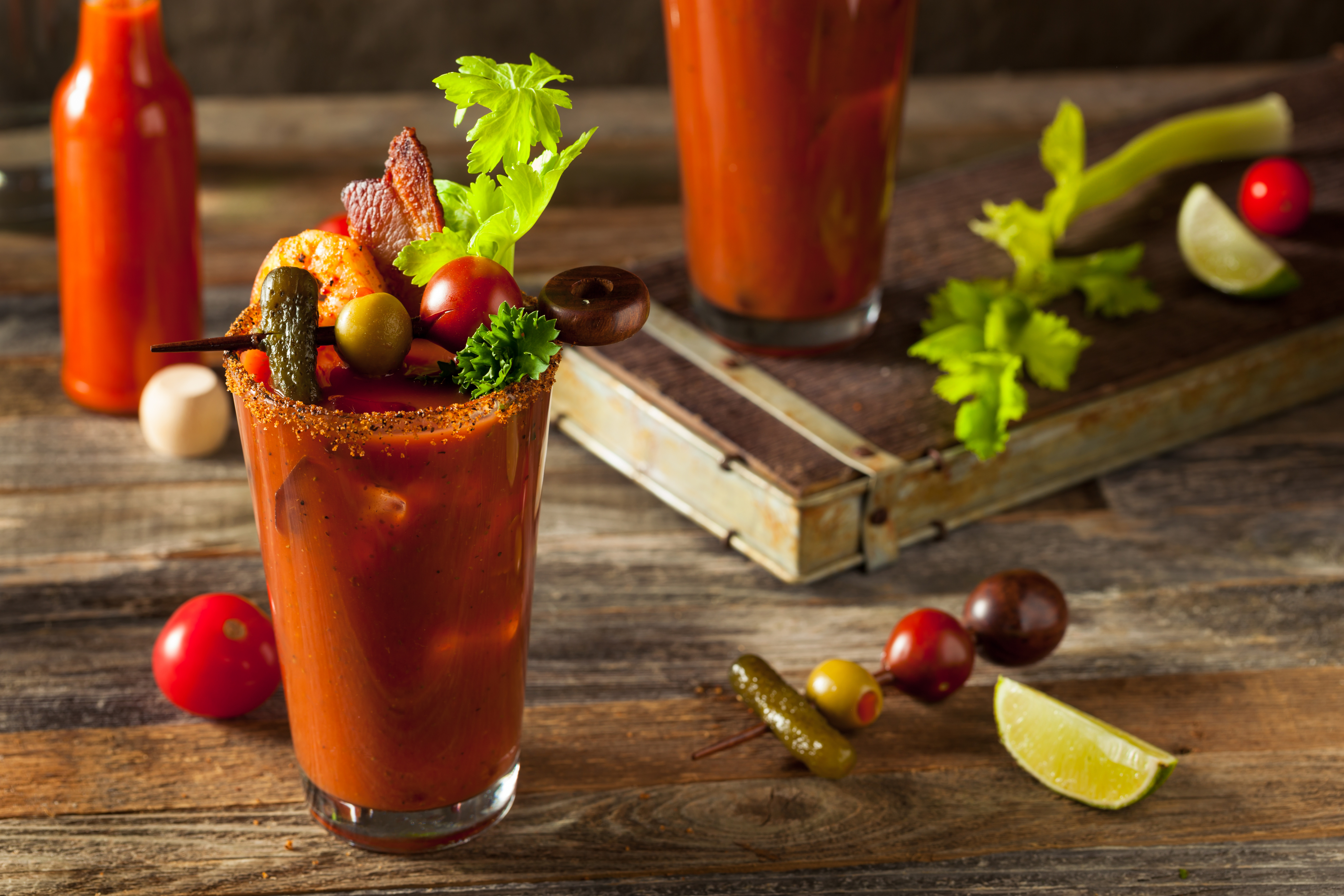 A glass of bloody mary with celery and other garnishes on a wooden table, with ingredients scattered around.