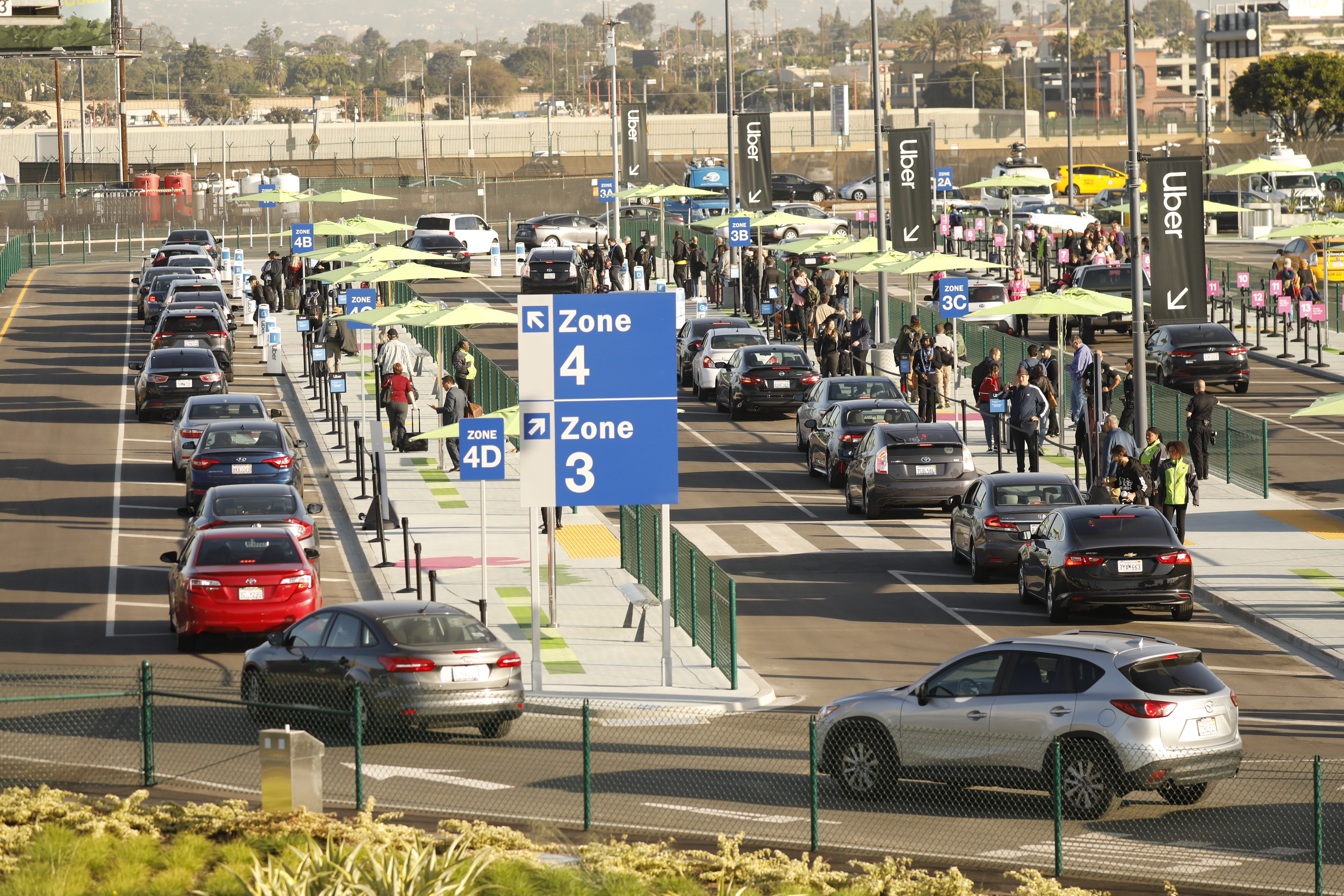 Rows of cars are lined up at an airport pickup parking lot with people holding luggage standing in long lines to access them. Signs that say Uber are seen in the background.