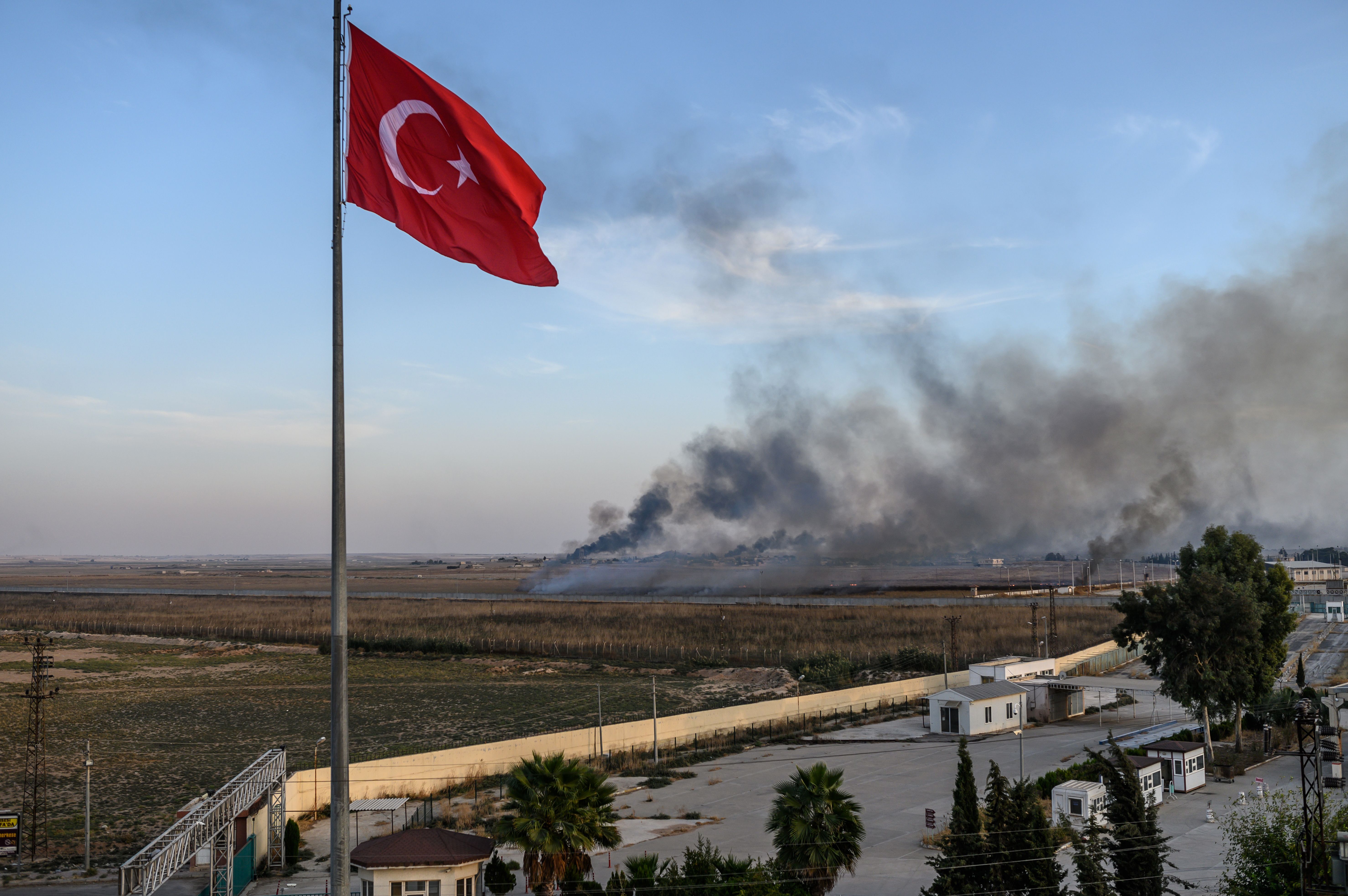 A Turkish flag flies in the foreground while smoke rises from over the border in Syria.