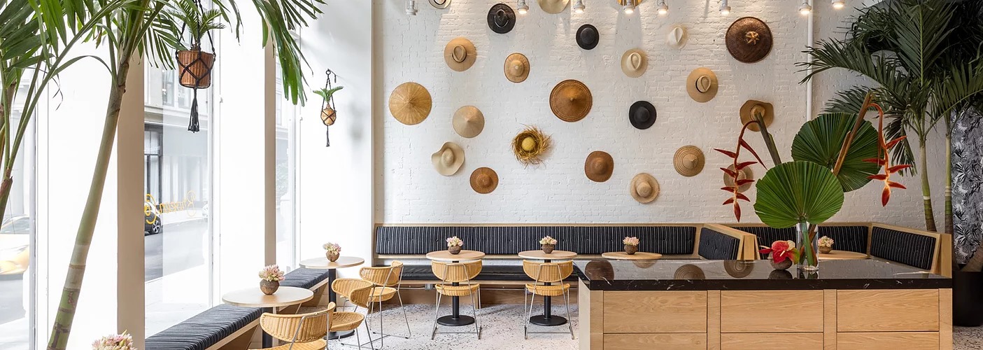 A bright white restaurant interior features palm trees and a wall of hanging beachy hats.