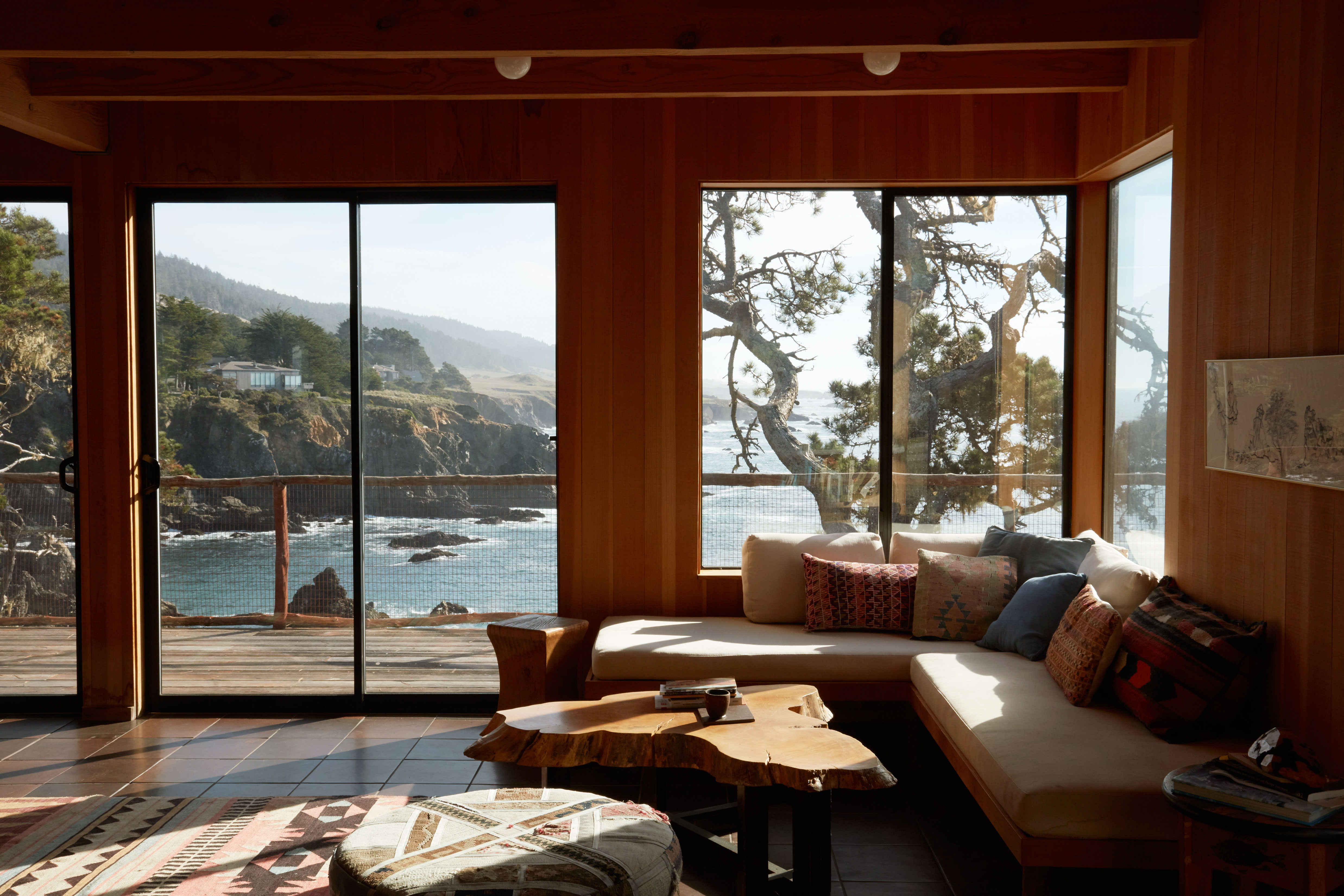 A living room overlooking the sea and some cliffs containers an L-shaped bench with 10 throw pillows. There is also a rustic, wooden coffee table and a geometric rug.