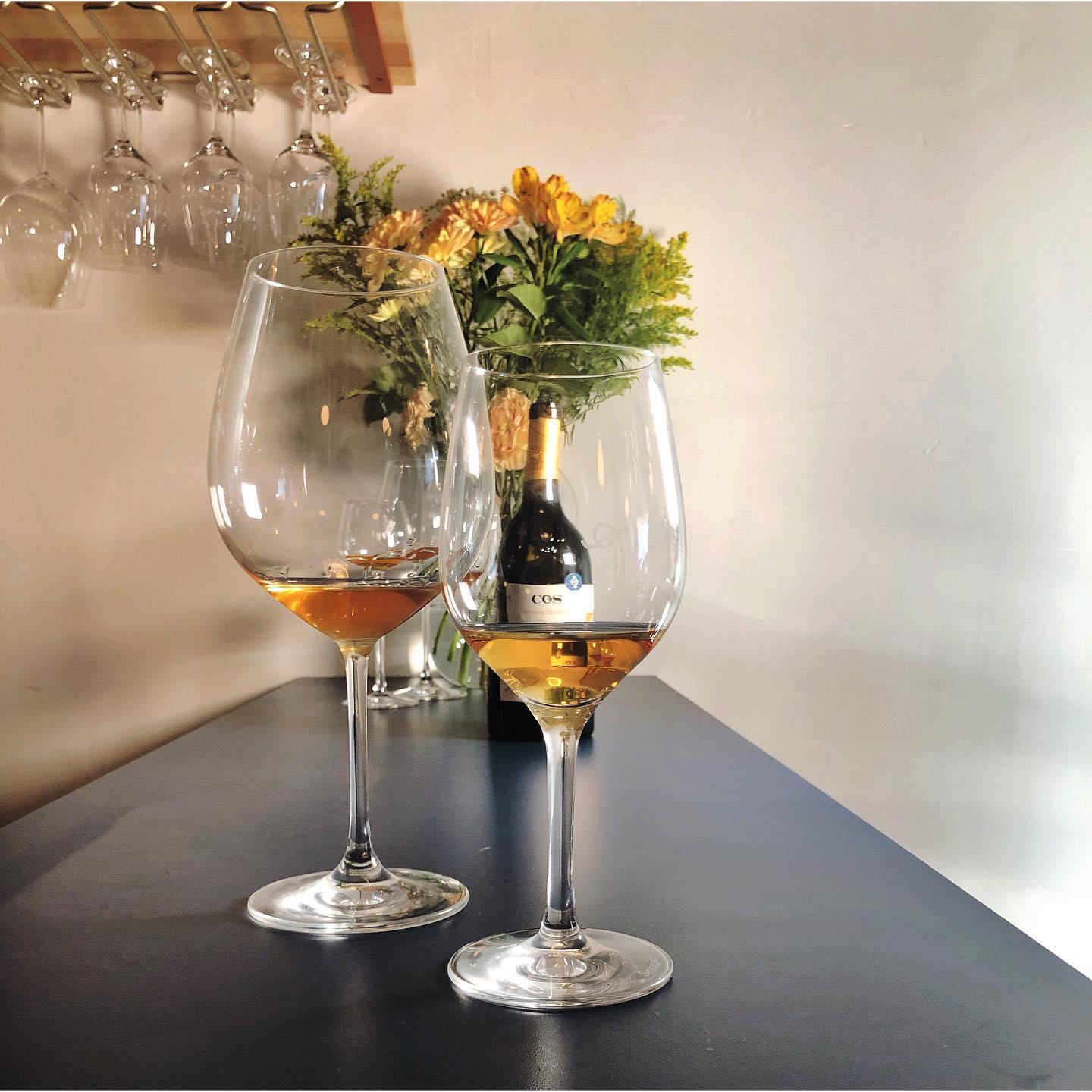 Two glasses of orange wine in front of a vase of flowers.