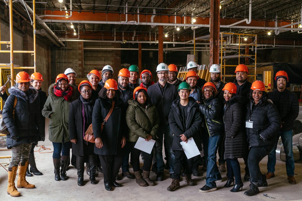 Two rows of people face the camera in a building mid-construction. They have on jackets and orange hard hats.