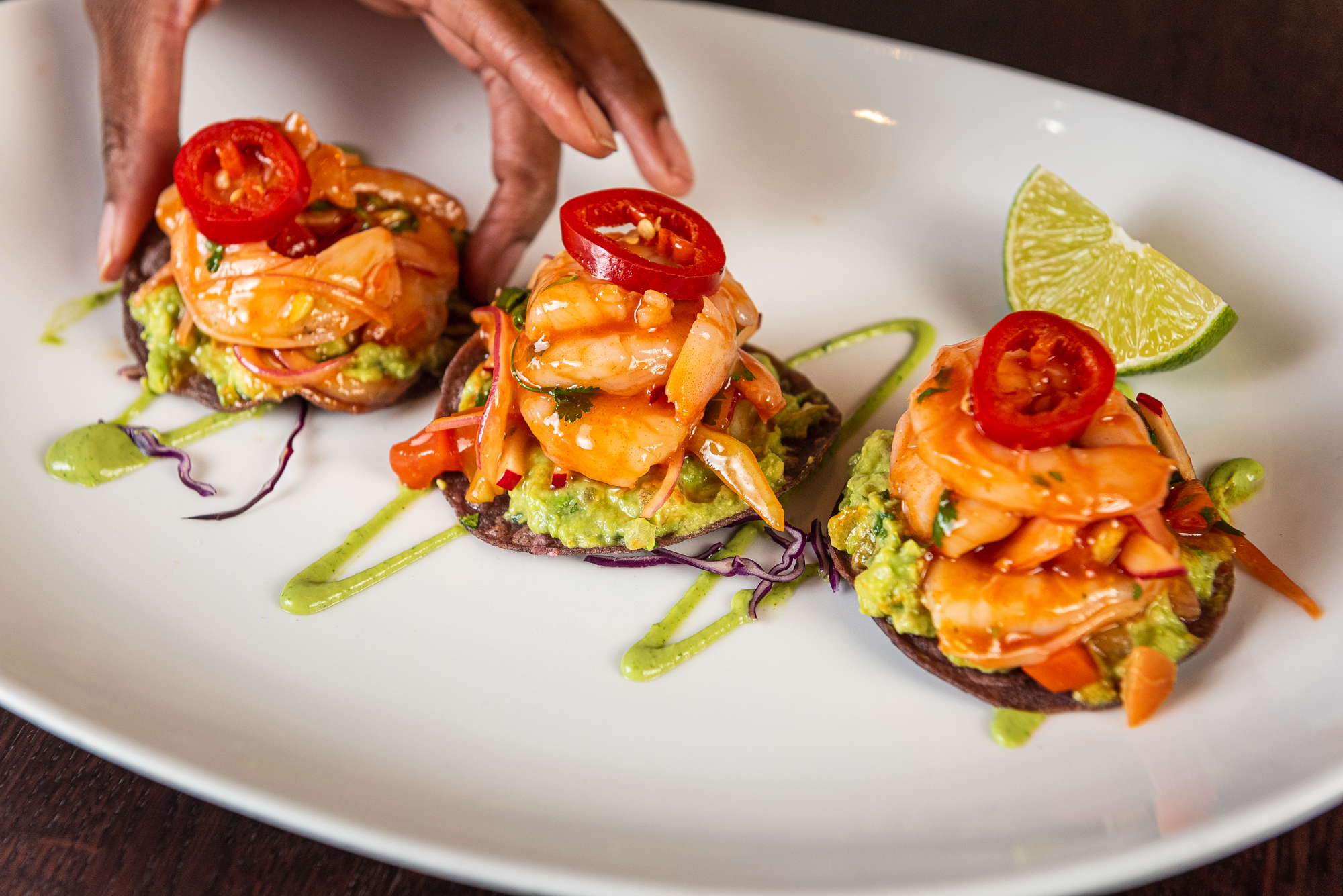 Three Yucatan shrimp tostadas at Anafre are built on blue tortillas with a base of green guacamole on a white plate.