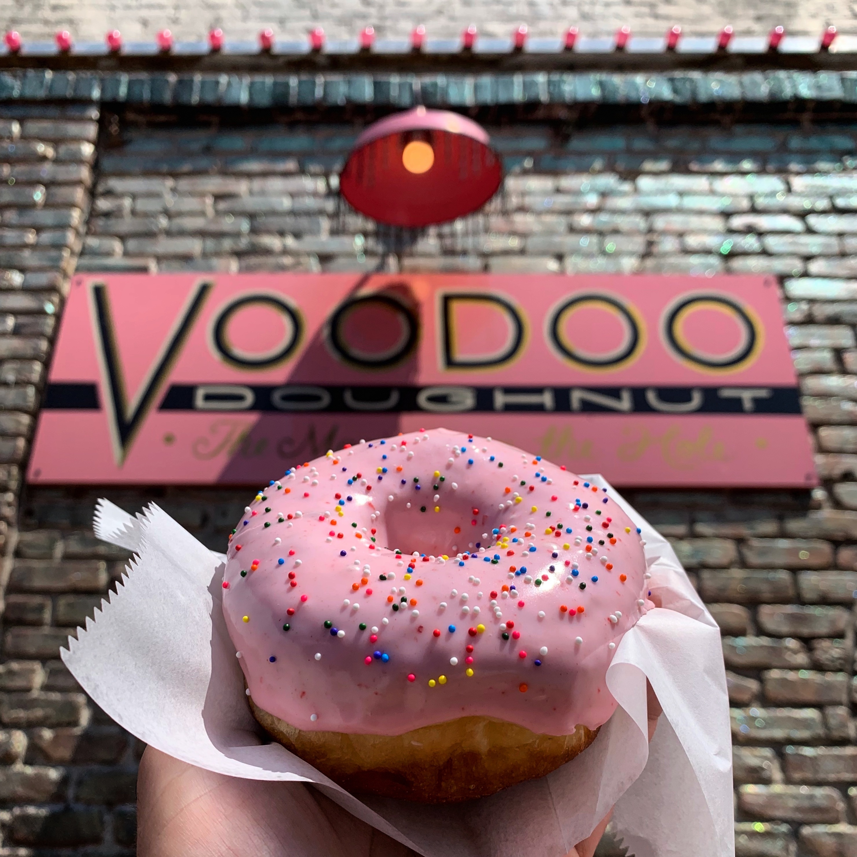 A picture of a hand holding up a Pepto-Bismol pink-glazed doughnut in front of the Voodoo Doughnut sign
