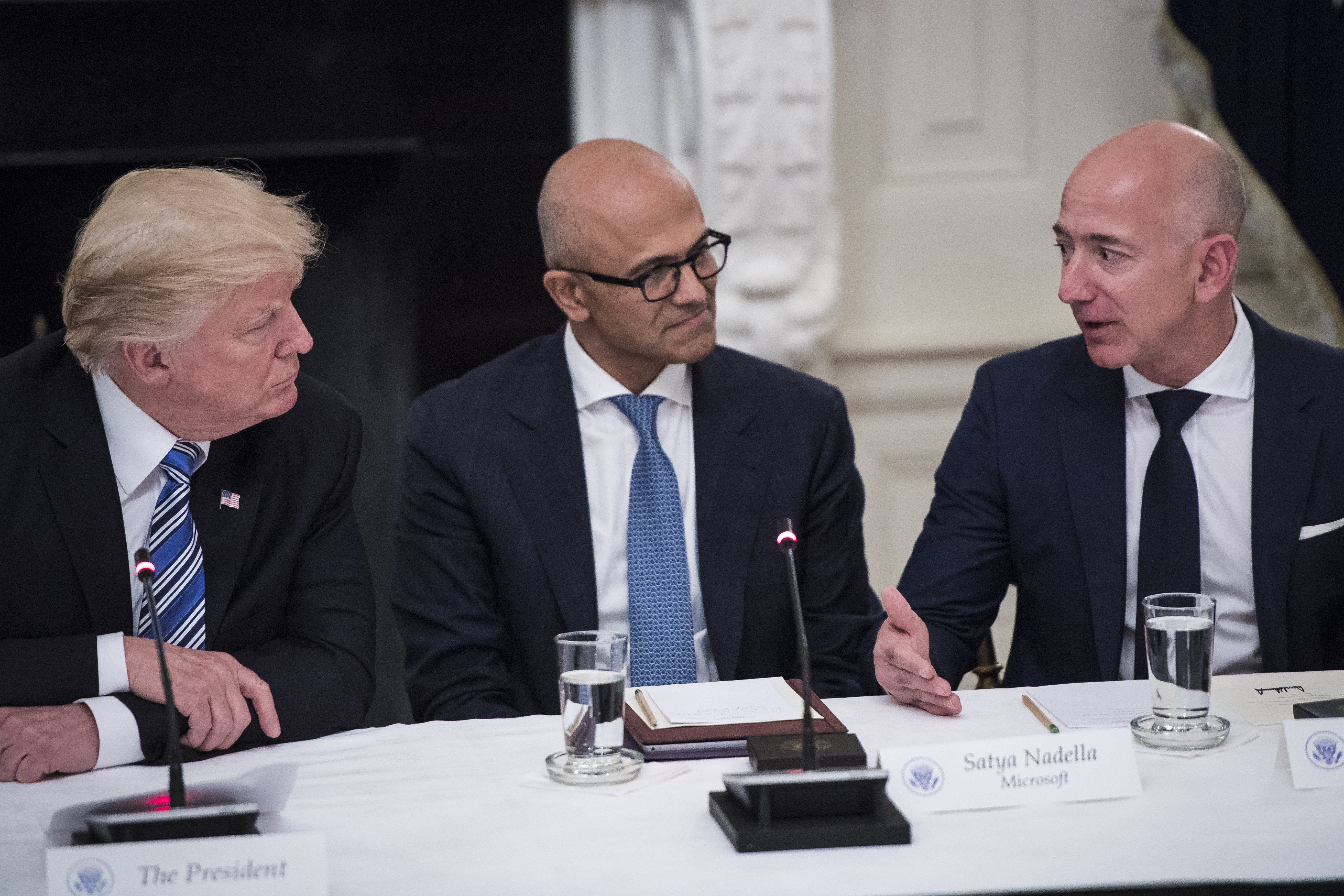 President Trump and Microsoft CEO Satya Nadella listen to Amazon CEO Jeff Bezos speak. All three are seated at a conference table.