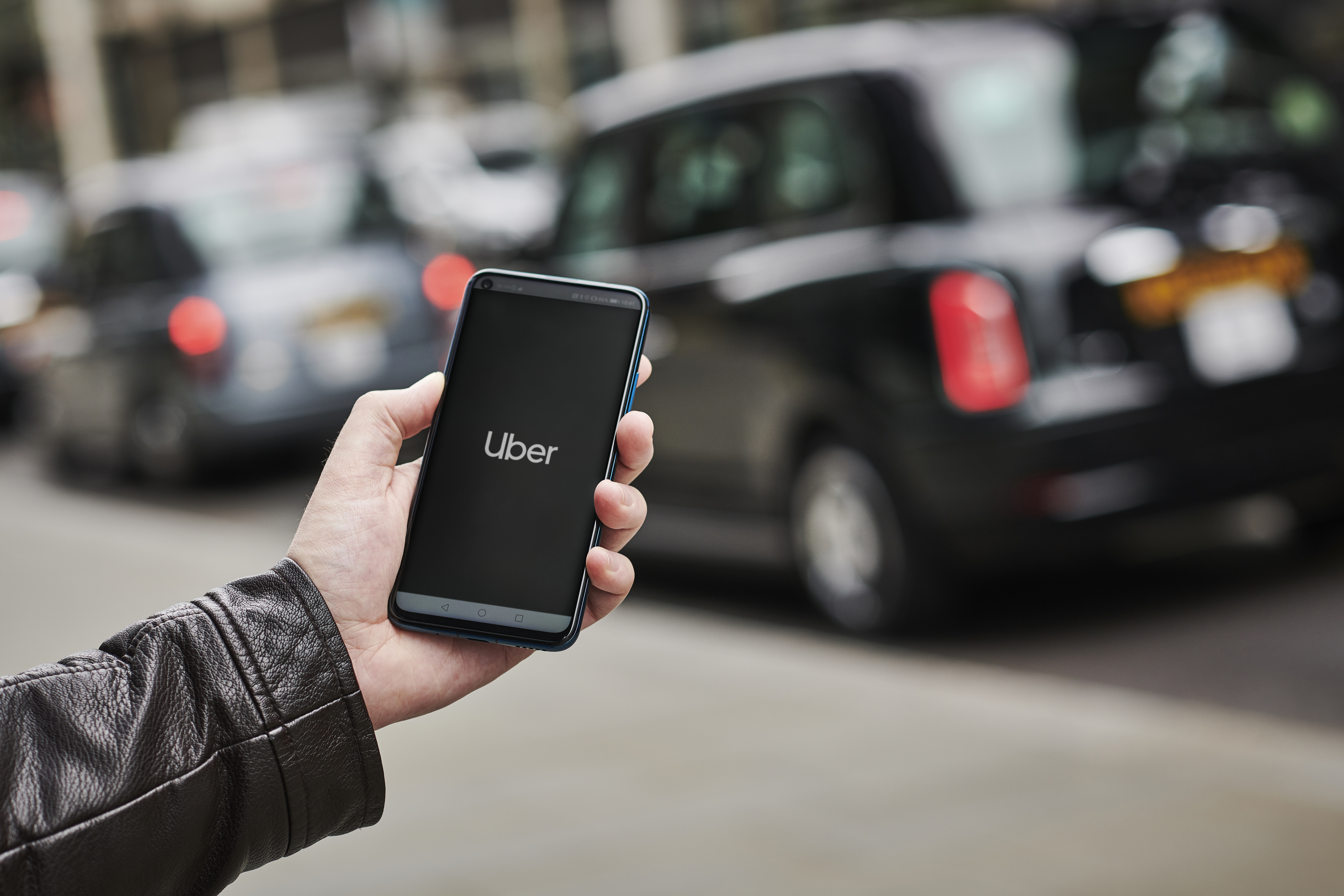 A man wearing a leather jacket holds a smartphone displaying the Uber app.