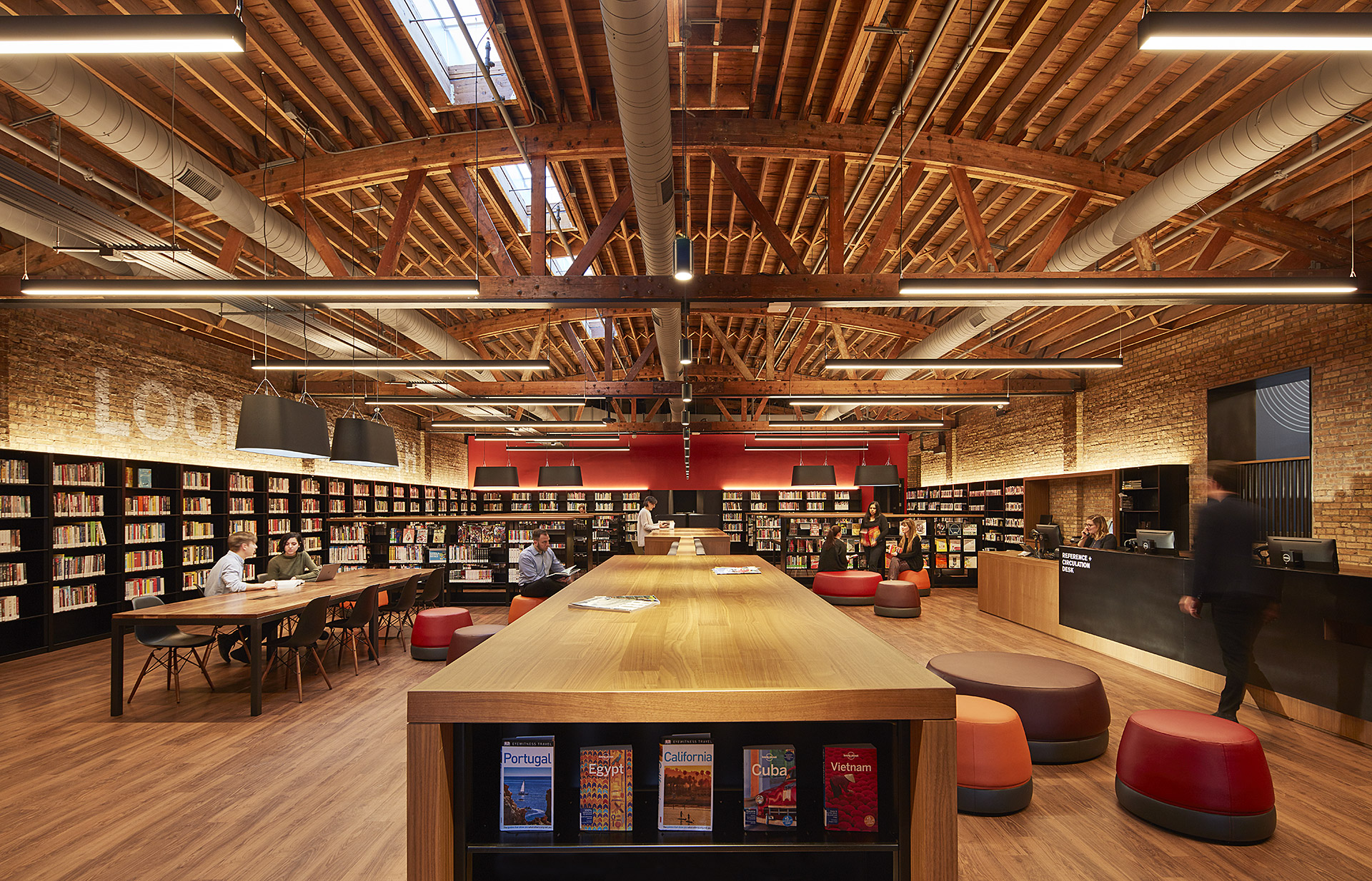 A long, rectangular library space beneath an arched wood timber ceiling. There are tables and chairs for reading the walls are lined with bookshelves. 