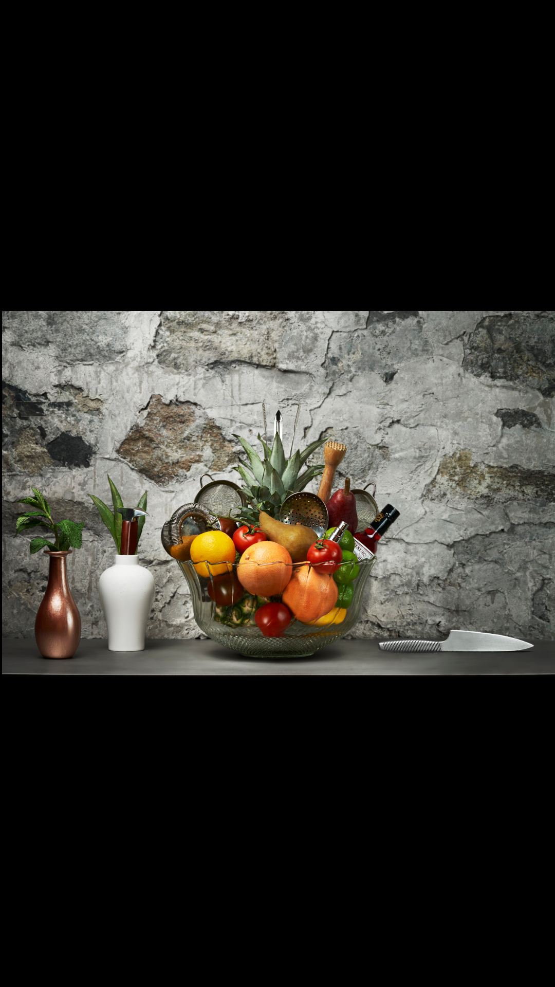 A glass bowl of fruit in front of a concrete wall.