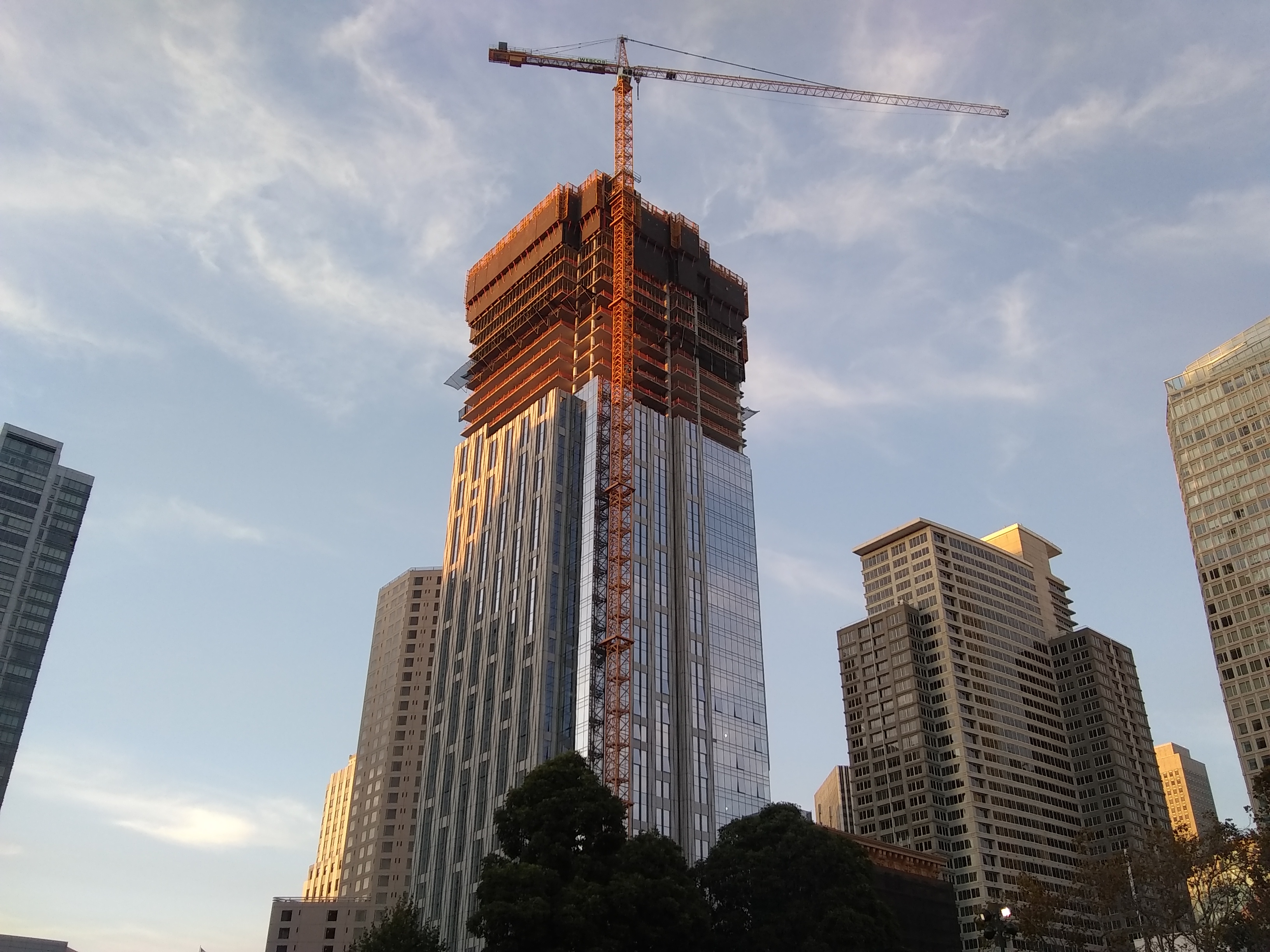 A construction crane perched on top of a partially built skyscraper, with glass installed on the bottom two-thirds of the facade.