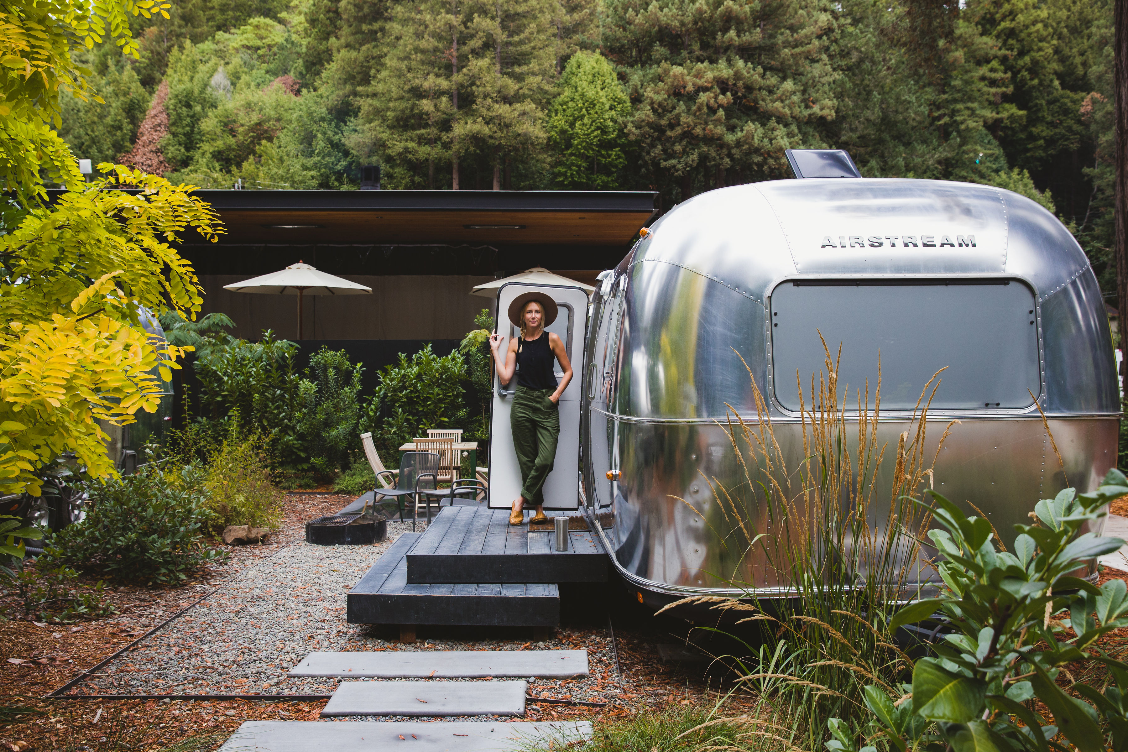 A silver bullet travel trailer by Airstream sits on a raised platform with a deck and landscaping. A woman stands in the trailer’s doorway. 