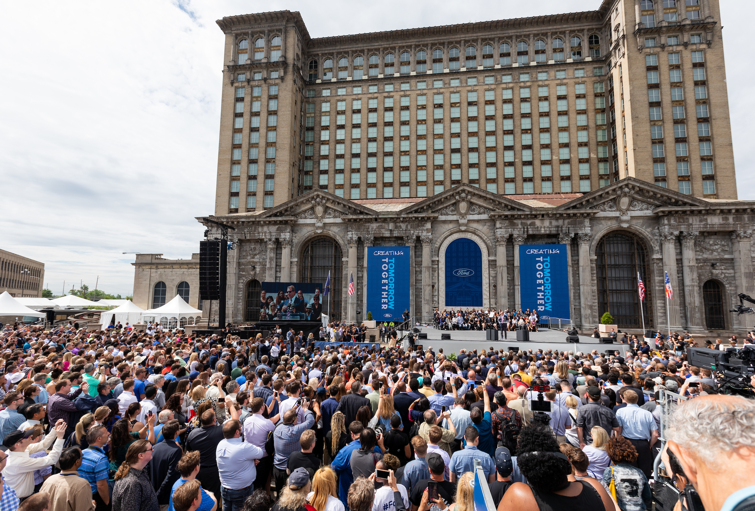 A large crowd of people gather outside a podium set up in the front of a tall, abandoned and ornate building. Two signs flanking the stage read “Creating Tomorrow Together.”