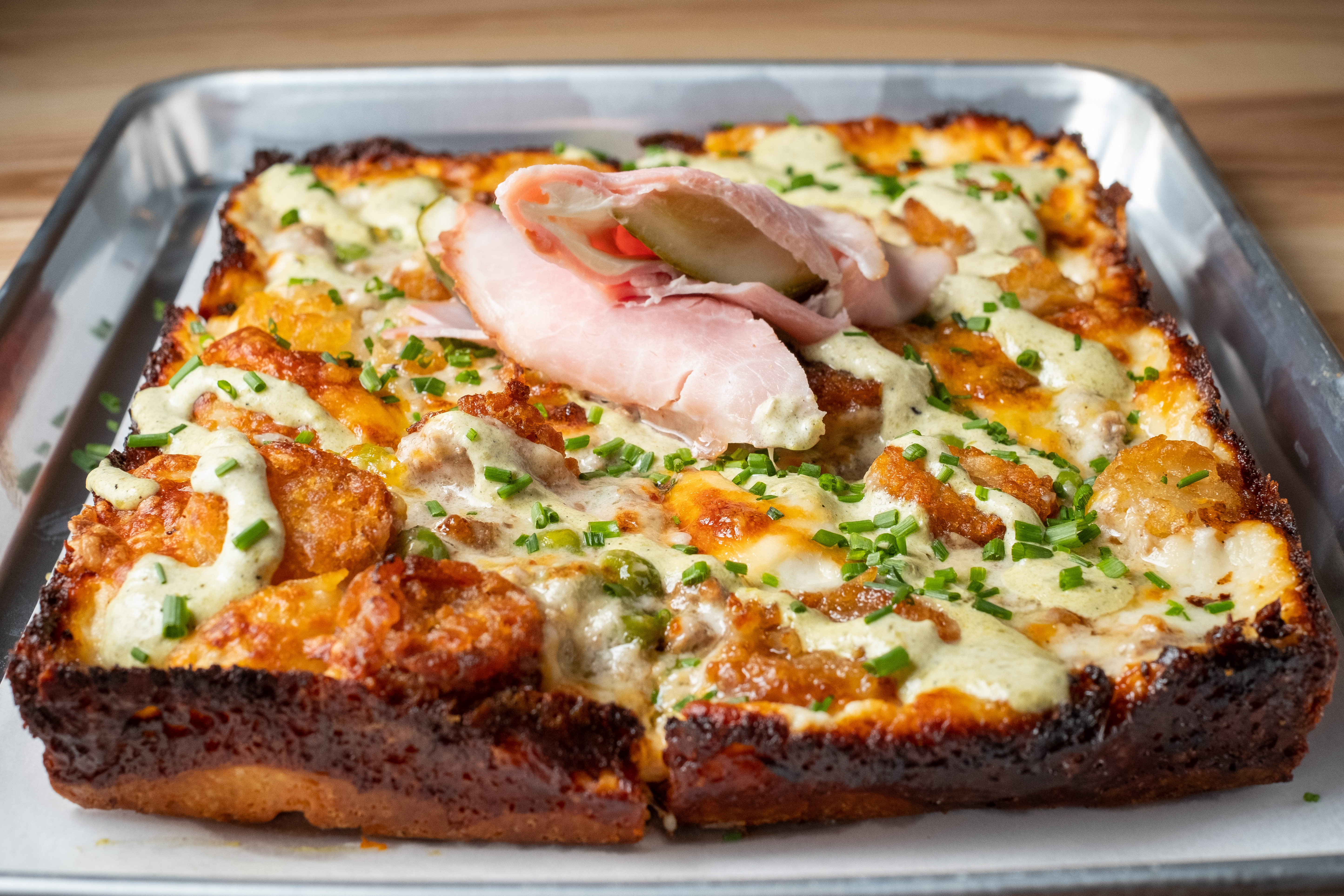 A rectangular pizza topped with tater tots, creamy sauce, an a pickle wrapped in ham garnish