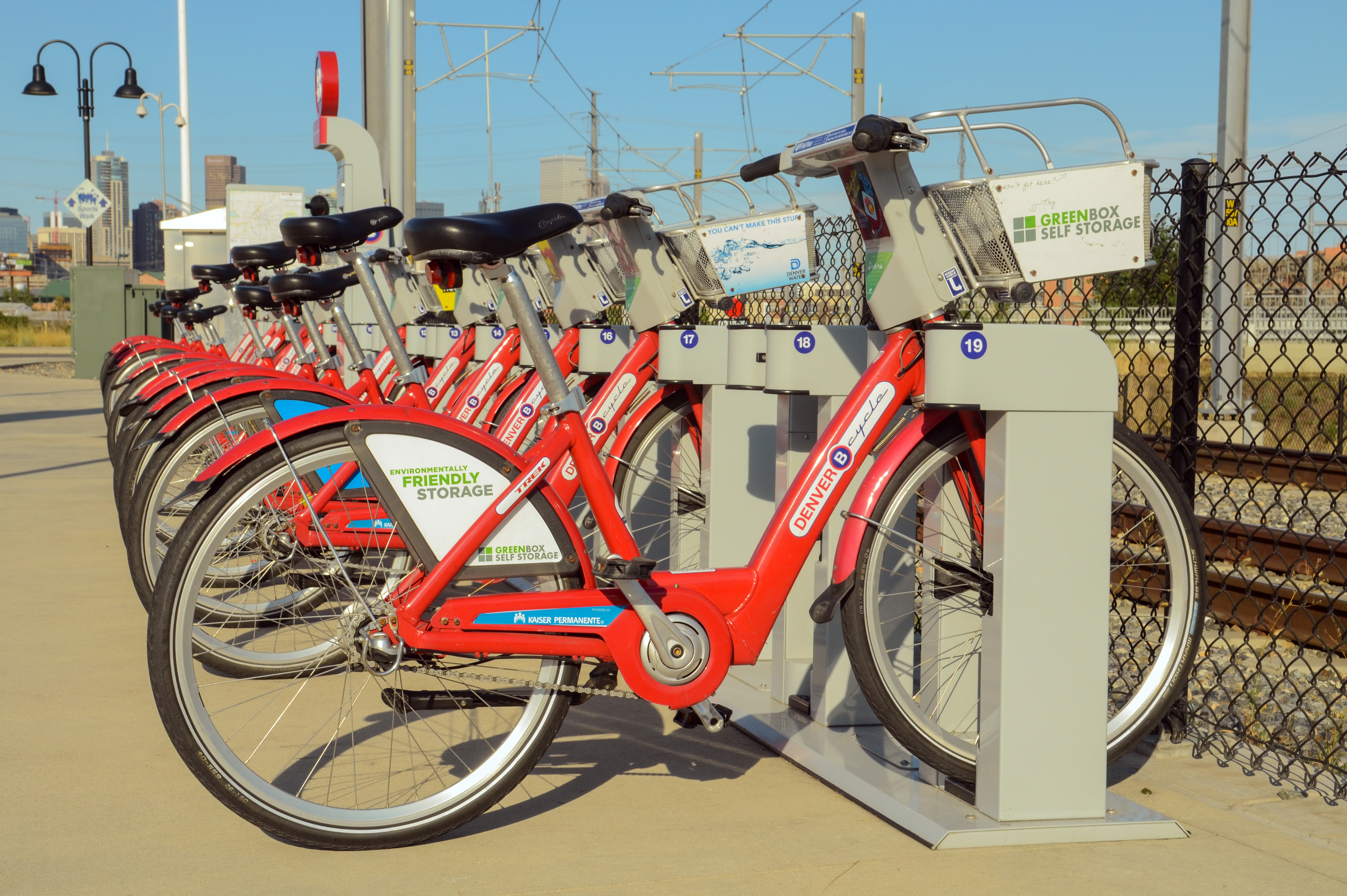 A row of bright red B-Cycle bikes parked at a bike-share station next to train tracks with a city skyline in the distance.