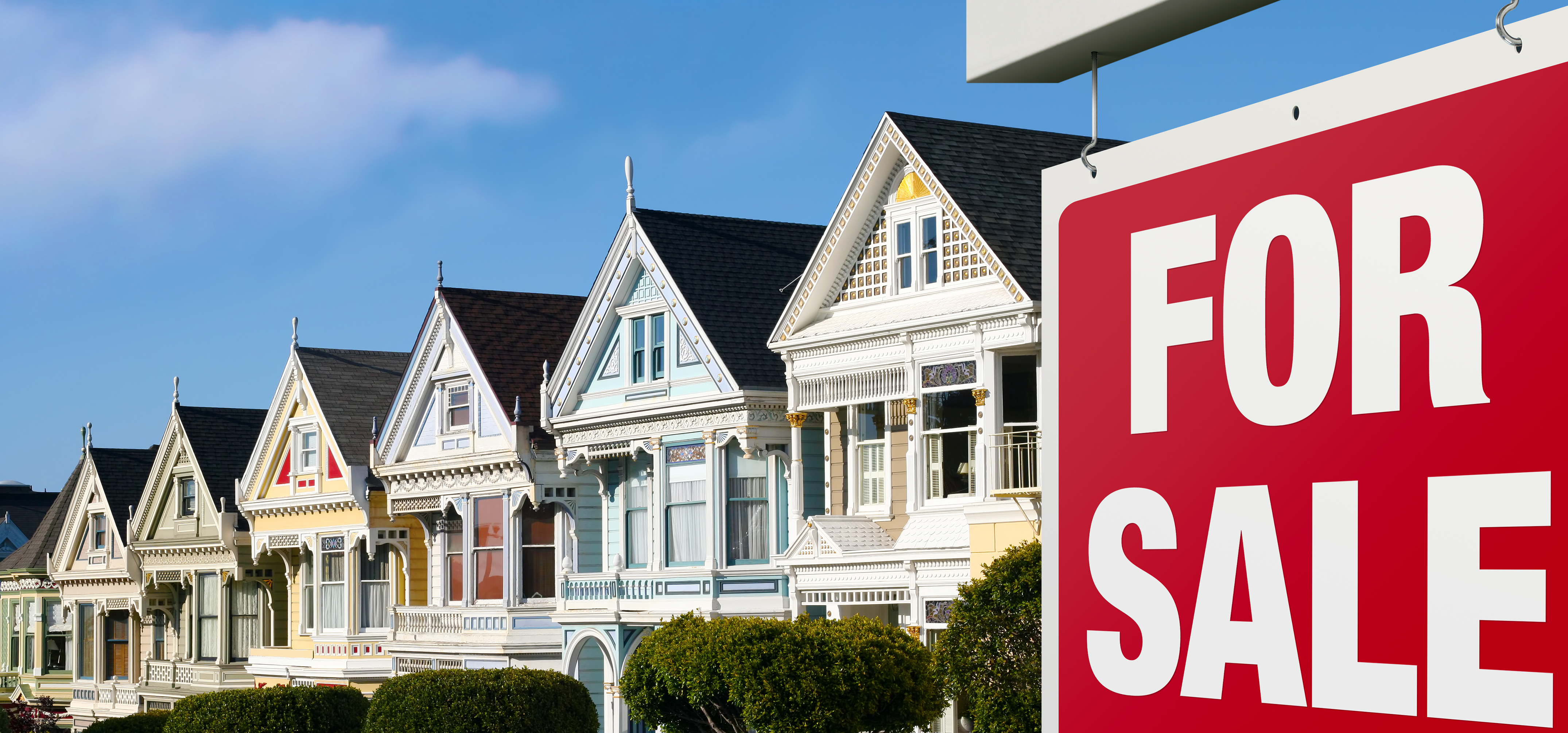 A row of colorful Victorian homes with a red and white “for sale” sign in font.