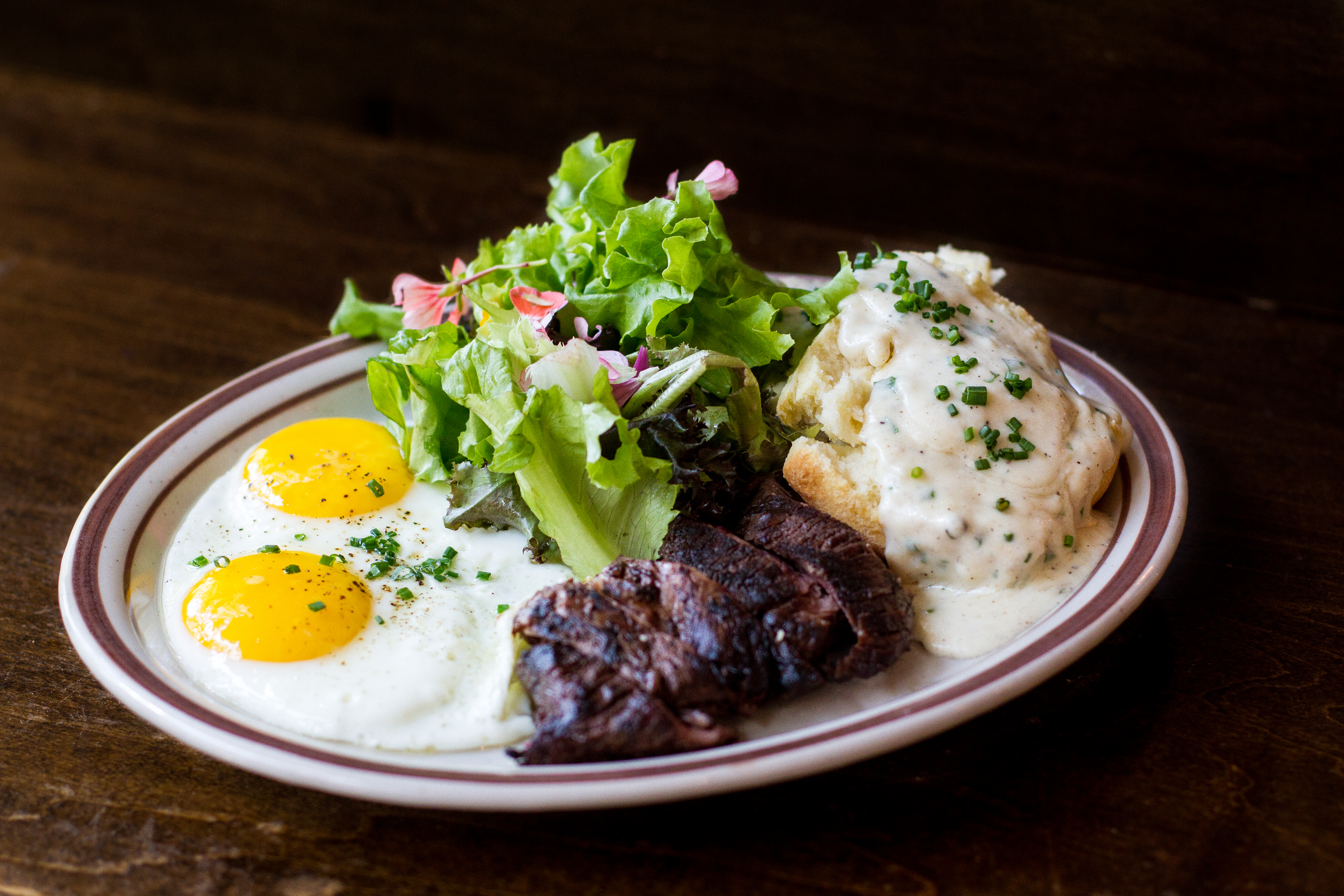 A white plate of two sunny side up eggs, a small streak filet, a biscuit with gravy, and a small salad
