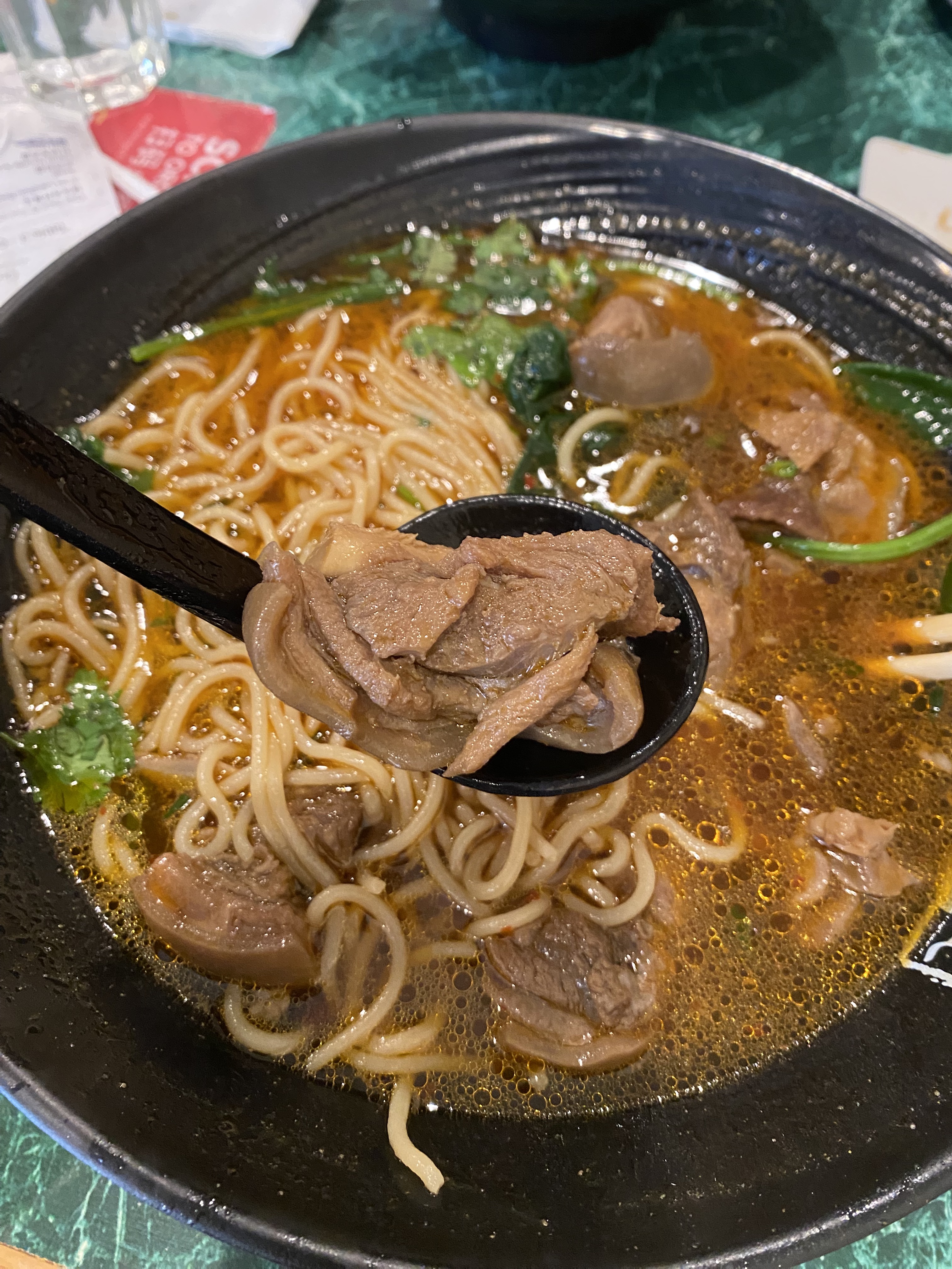 A bowl of noodles in broth with a spoon held up to display pieces of meat included in the soup.