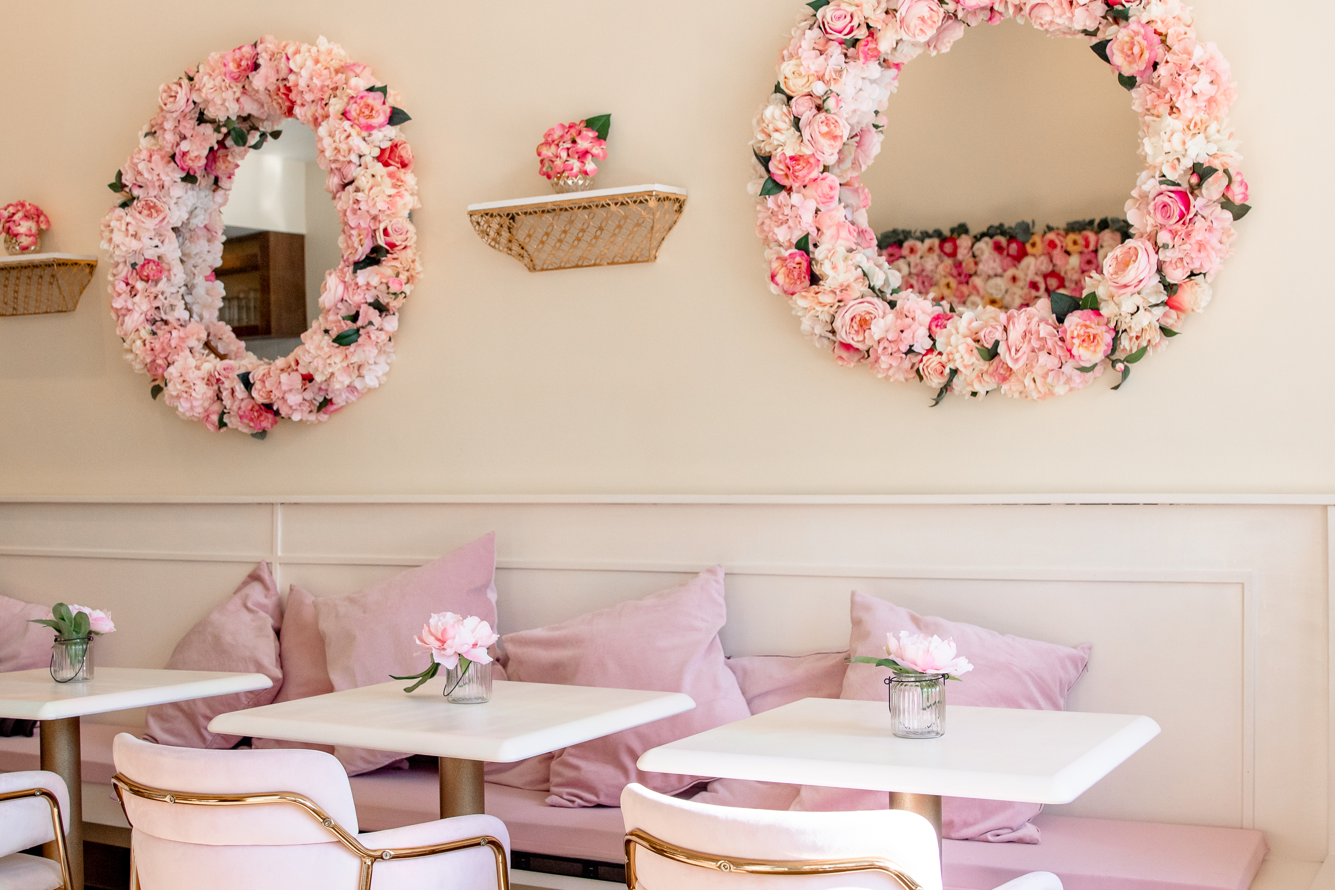 The creamy walls are offset by pale pink pillows on a pink velvet bench, and white tables are flanked by pale pink velvet and gold chairs