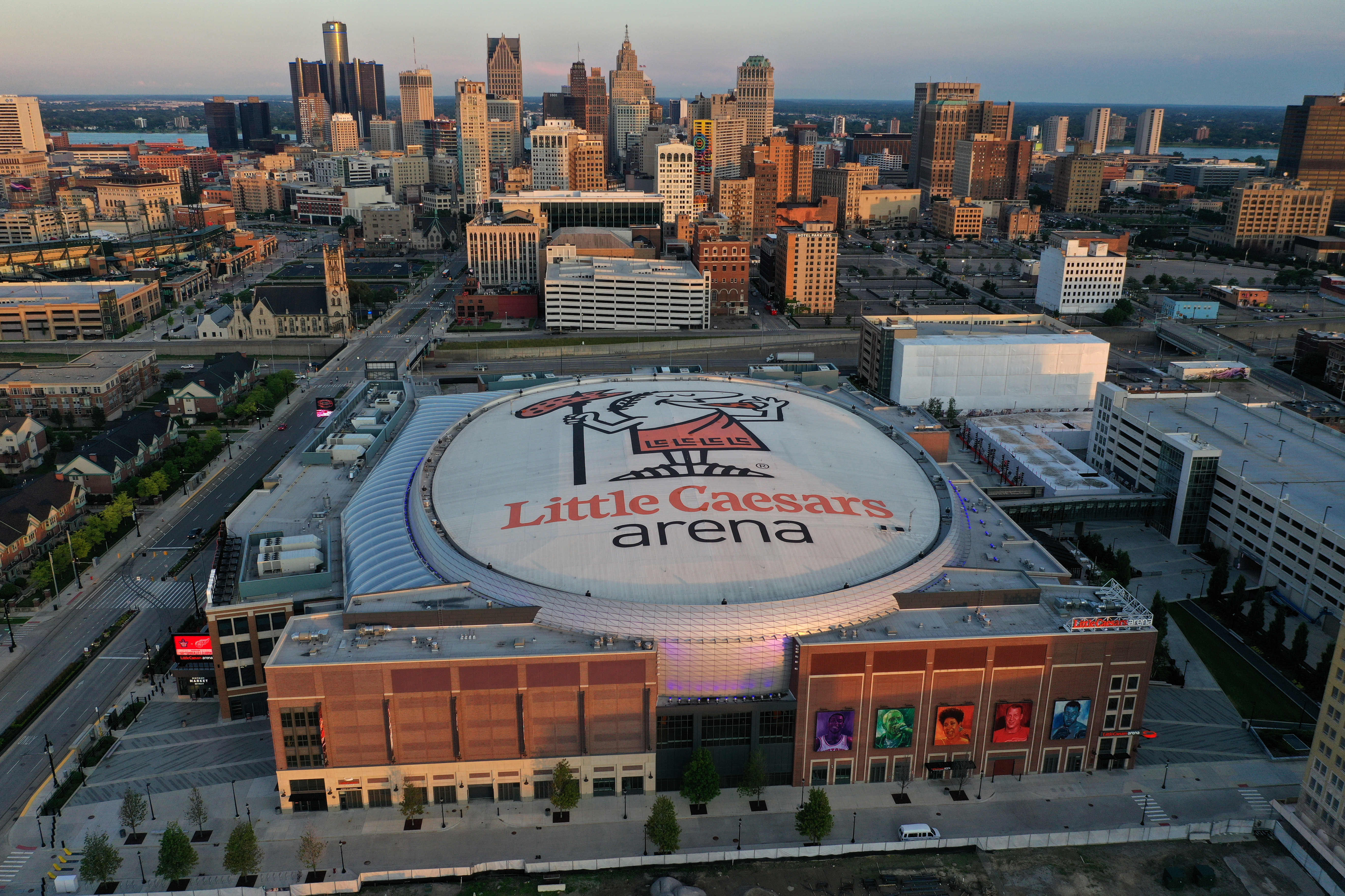 Aerial view of a huge arena with Little Caesars Arena on the dome. Many tall buildings are nearby.