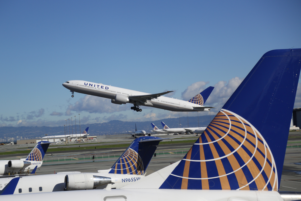 An airliner taking off, with the large tail of a different plane in the foreground.