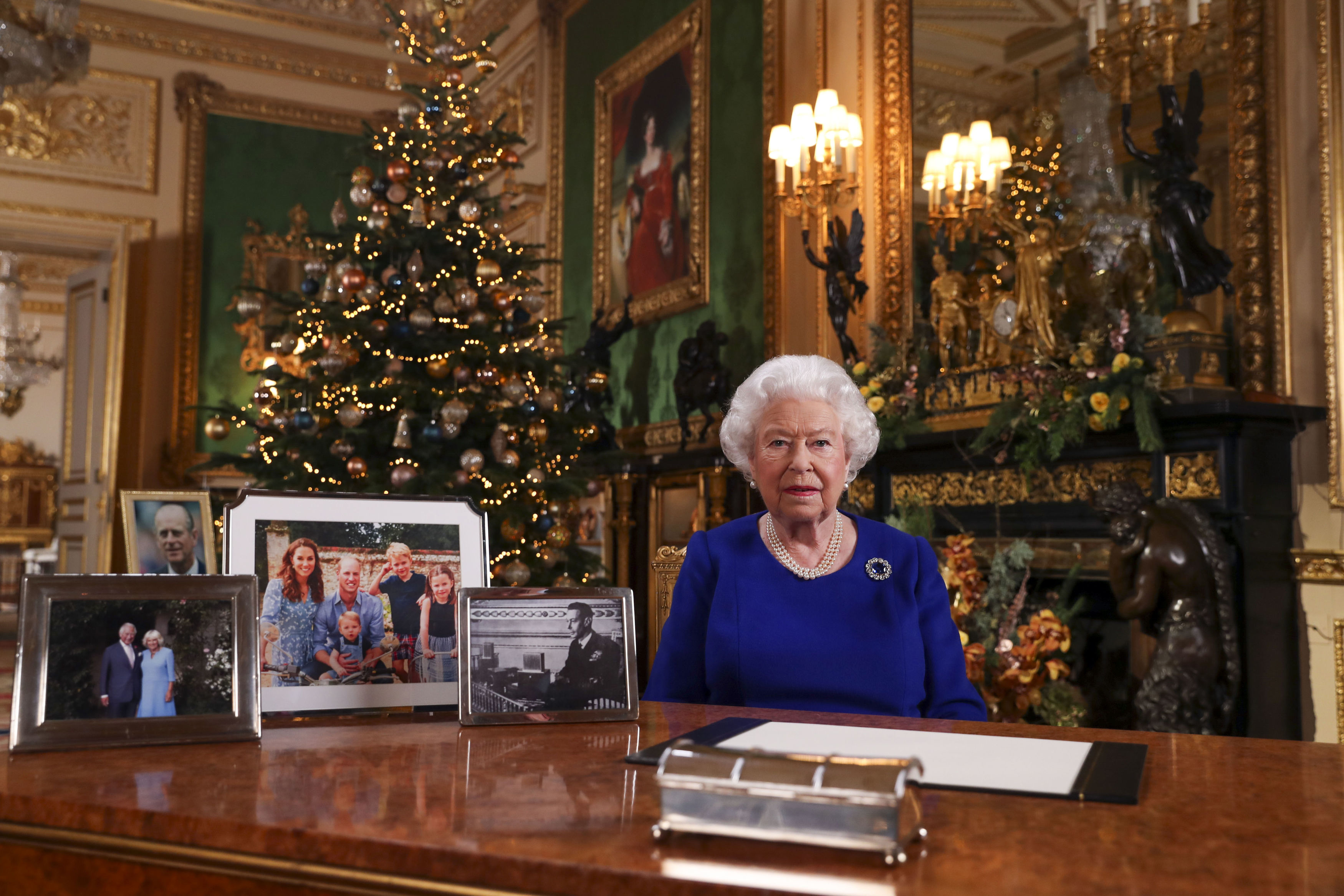The Queen of England sitting at a table with a Christmas tree behind her.
