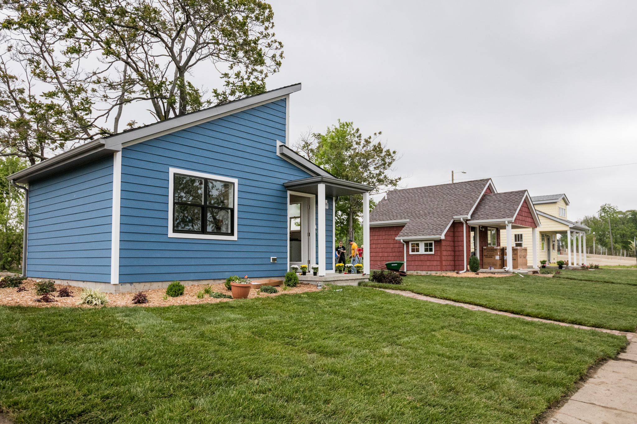 Three small single-story homes, colored blue, red, and white, sit in a row behind well-mowed lawns.