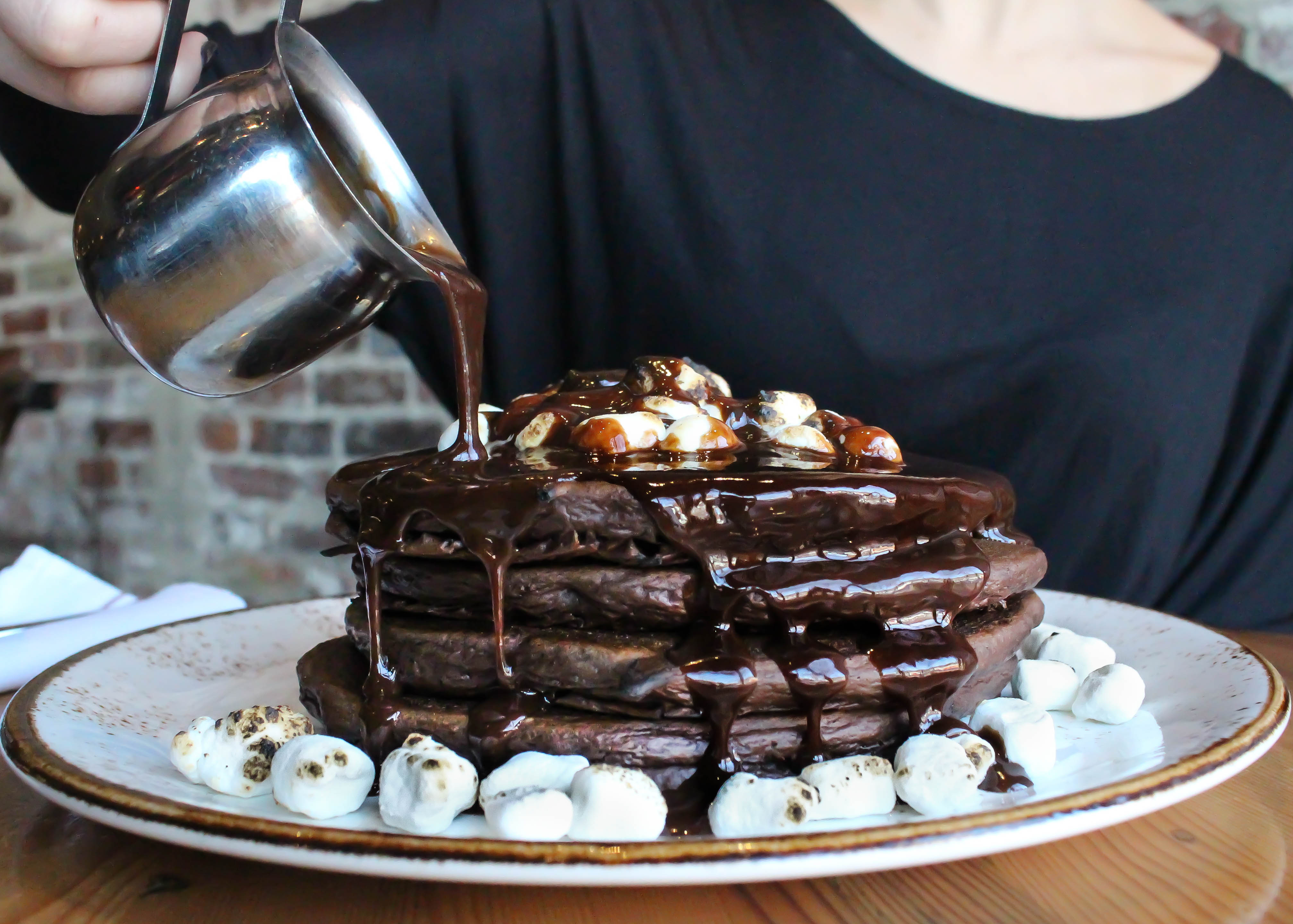 A stack of dark brown pancakes - made with hot chocolate - is topped with marshmallows. A person whose face isn’t visible in the photo pours a metal container of chocolate sauce over the stack.