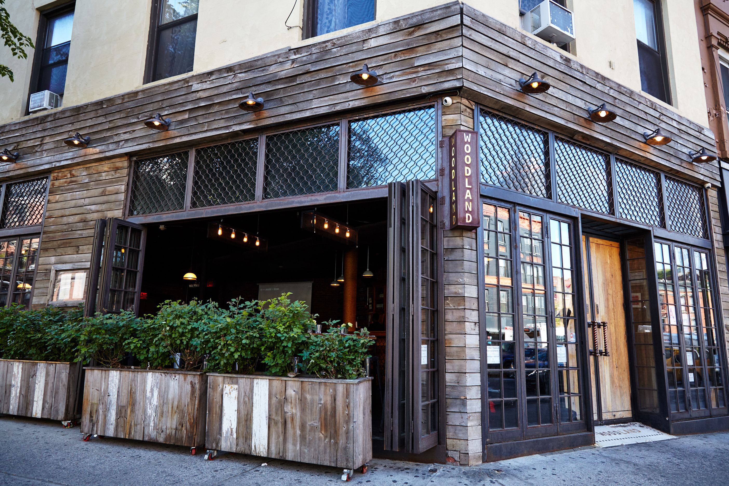 The exterior of woodland bar in park slope shows dark wooden frames, large glass windows and doors, and planters