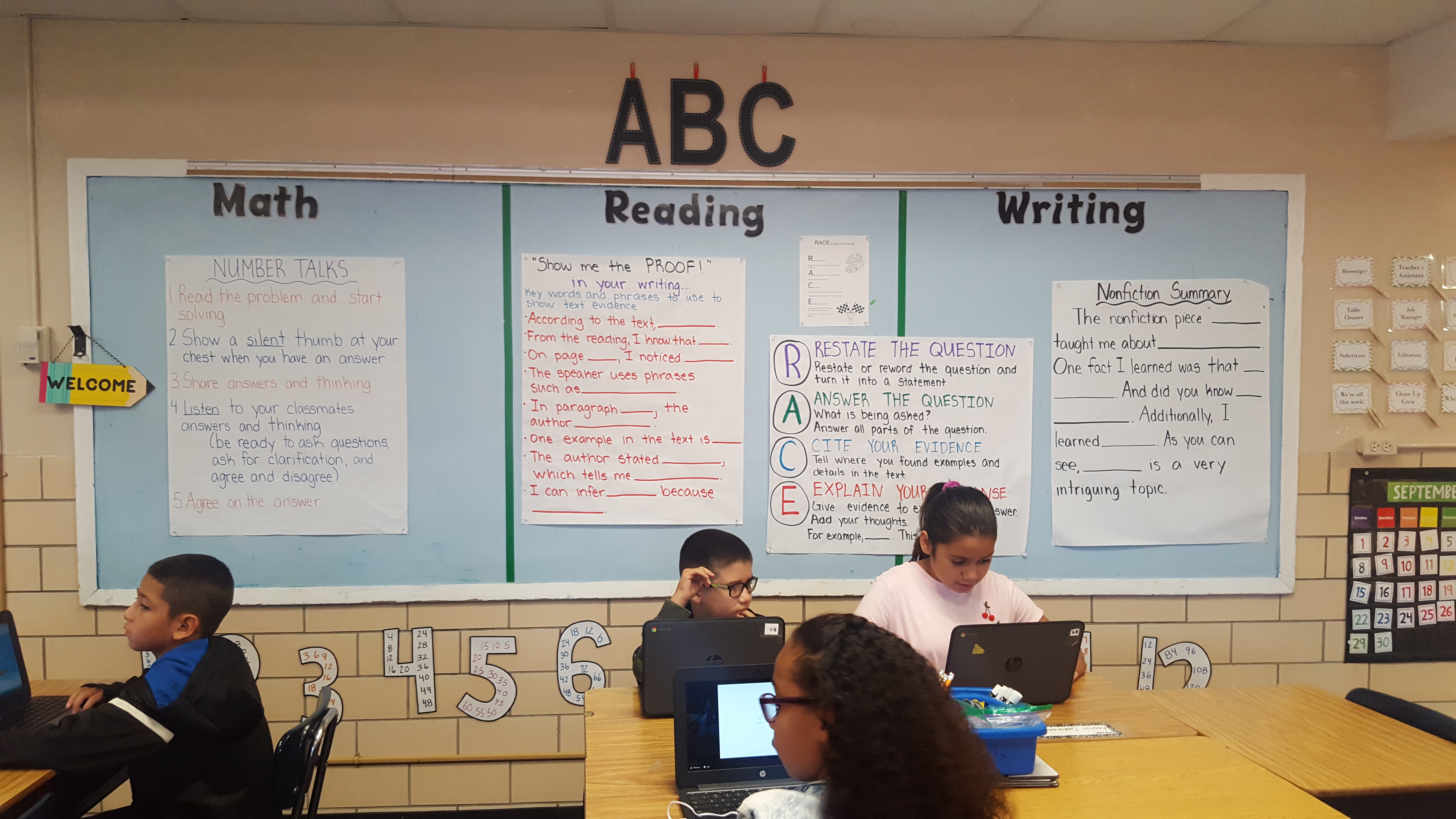Elementary students work at their desks. The wall behind them is decorated with math, reading, and writing lessons. Large letters ABC are above.