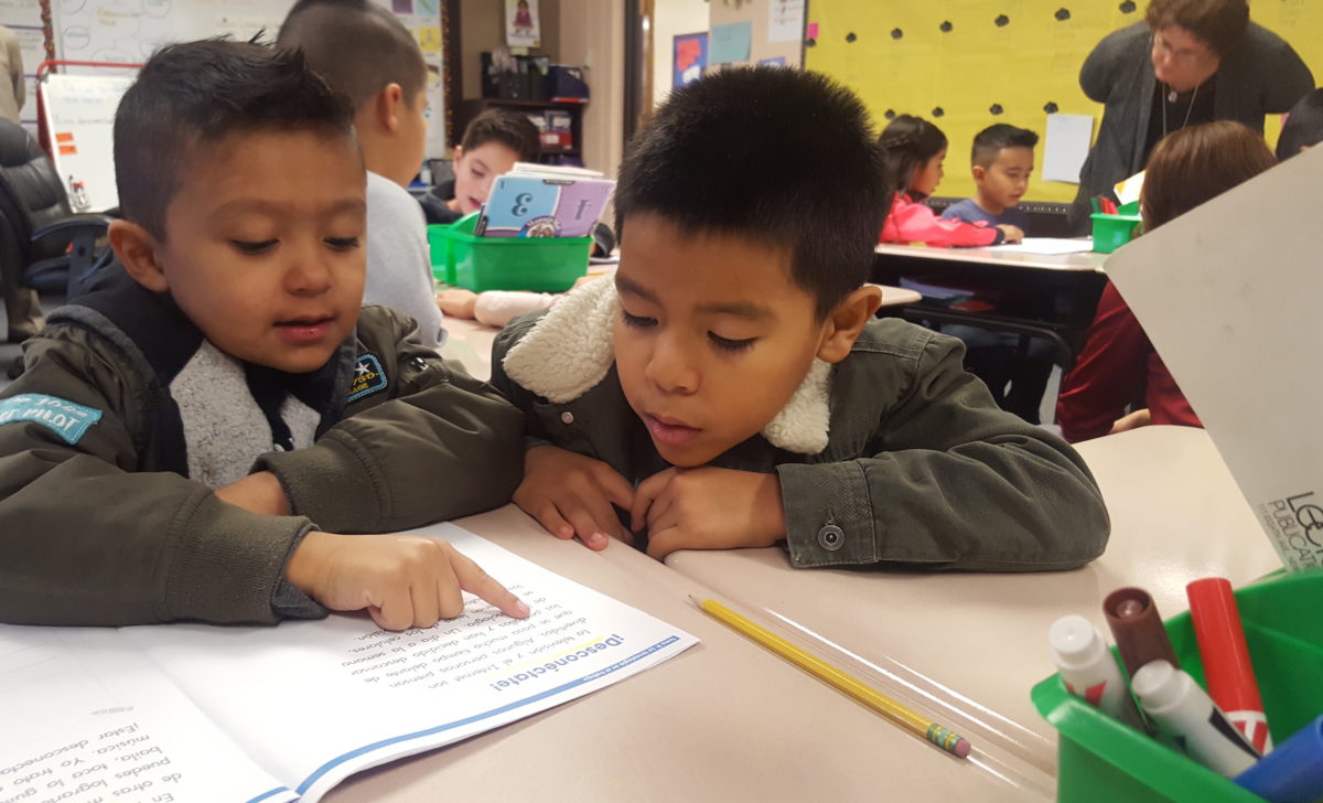 Two first grade boys look at a reading workbook at a desk in a classroom.
