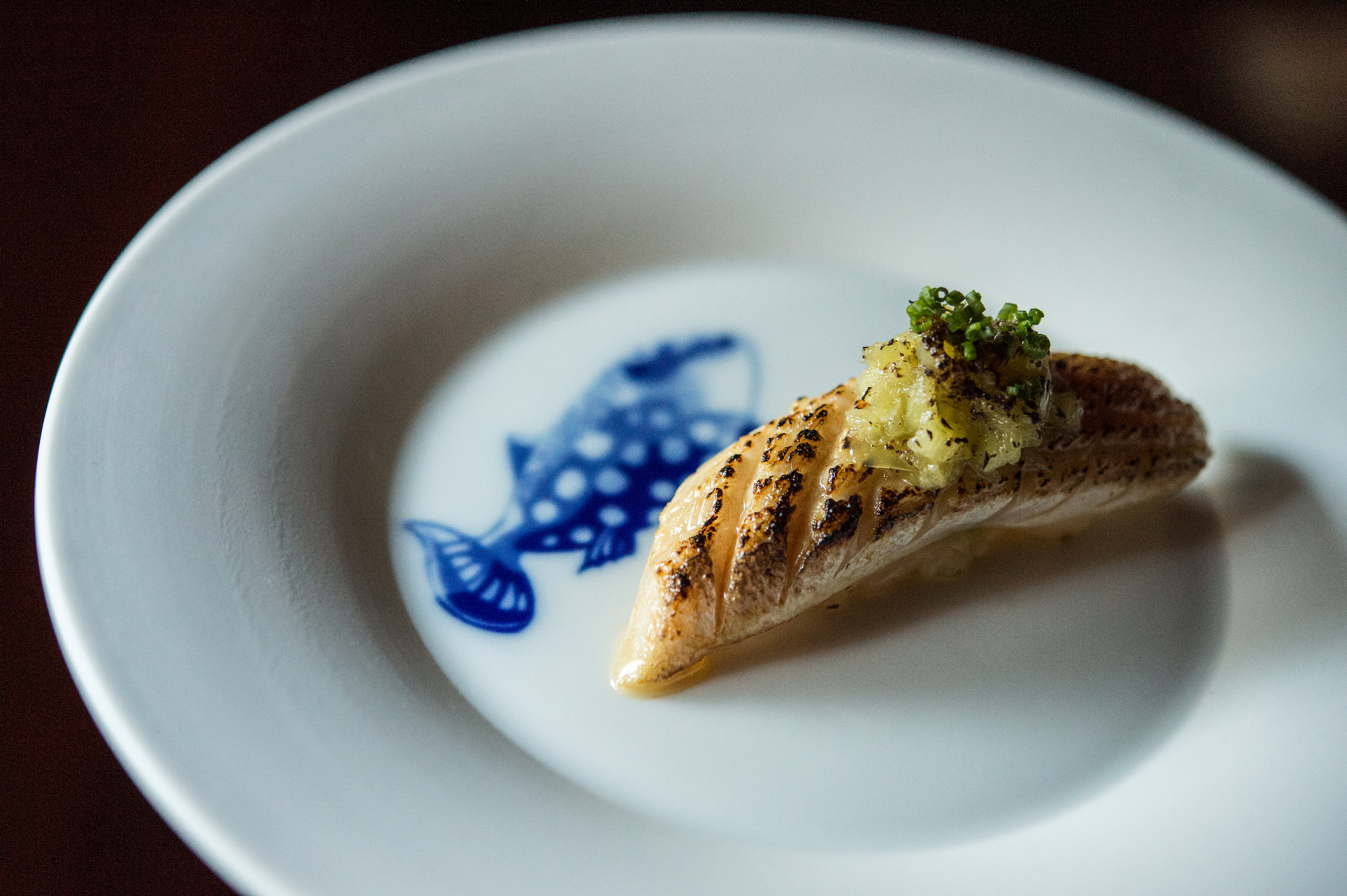 A white plate with a blue fish illustration holds a single piece if hamachi with banana pepper mousse.