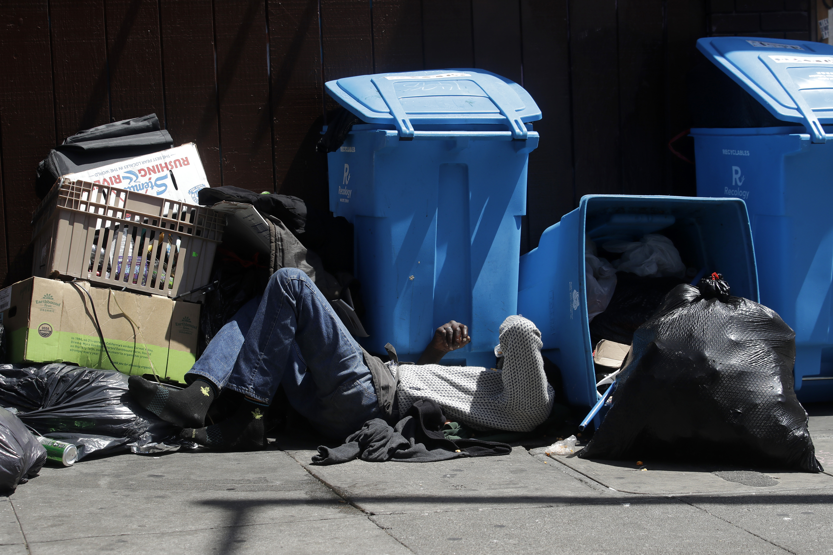 A blue recycling bin and a full black garbage bag on a sidewalk with a man sleeping next to them as he shields his eyes from the sun.