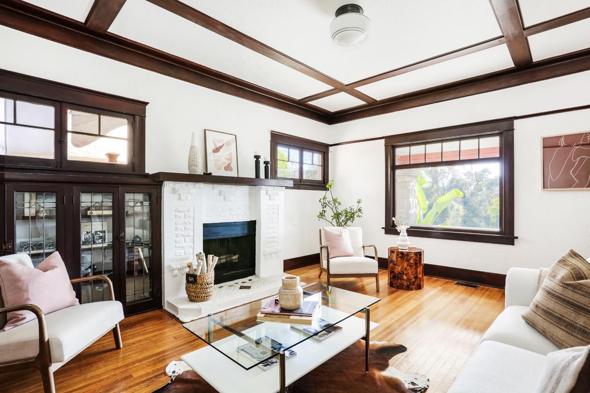 A living room with extensive woodwork and a brick fireplace that has been painted white. The room is furnished with a cowhide rug, glass coffee table, and white sofa. 