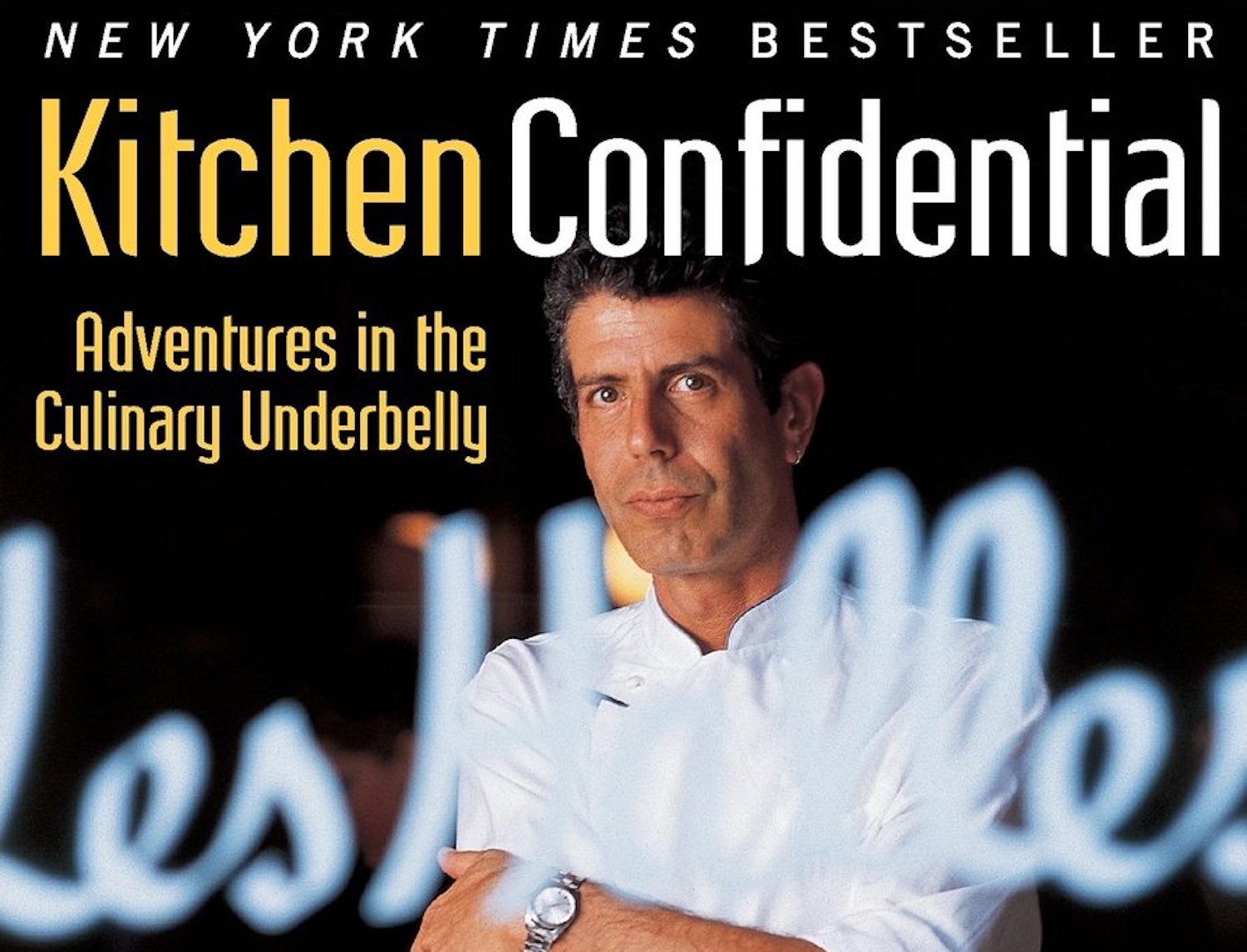 On the book cover for Kitchen Confidential, Anthony Bourdain, with salt and pepper hair, stands dressed in a buttoned-up chef’s coat a in front of restaurant windows painted with the words “les halles.” The book’s title appears above his head in bold yellow block letters.