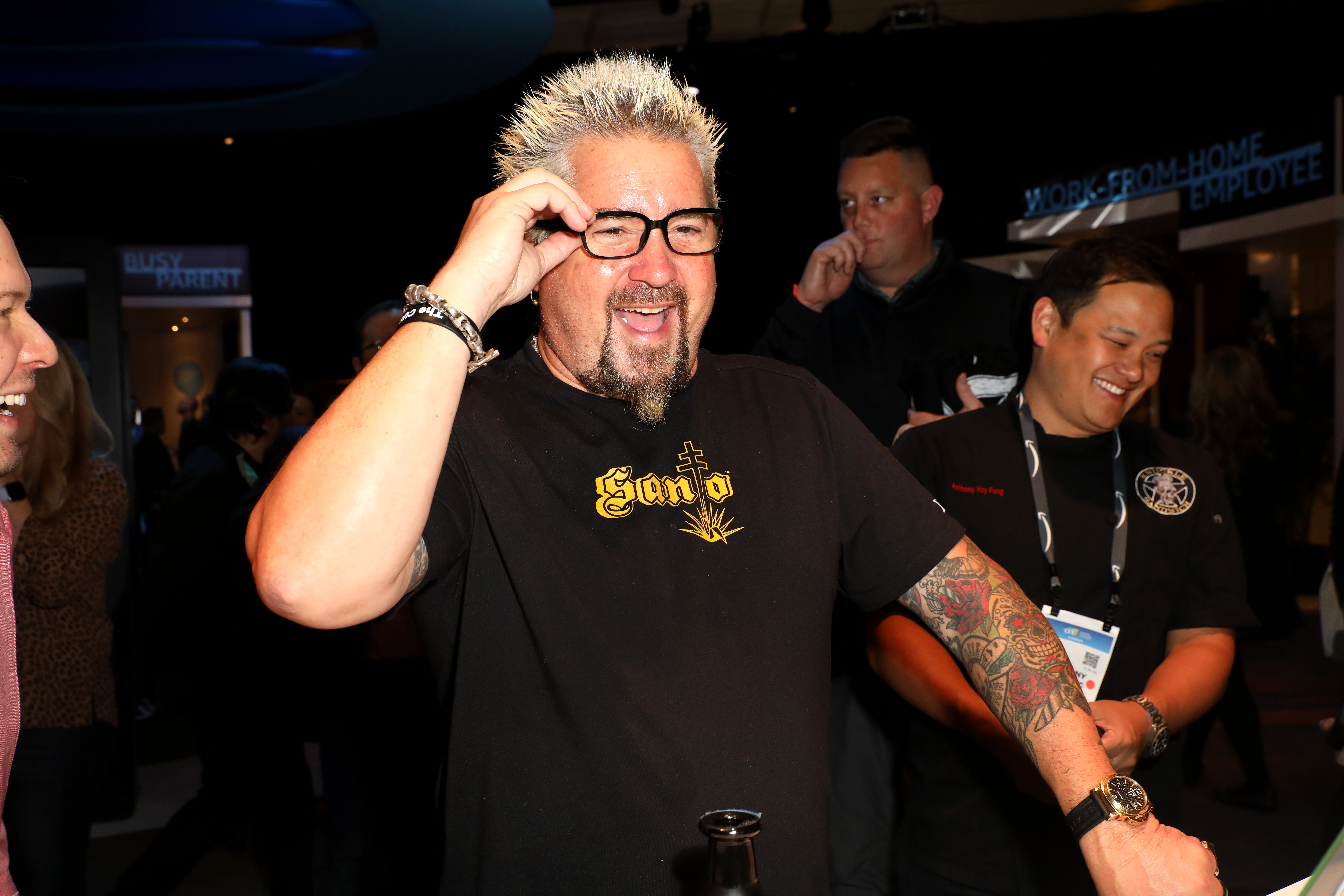 Guy Fieri, wearing a black t-shirt and glasses, walks by with his famously spiky blond hair