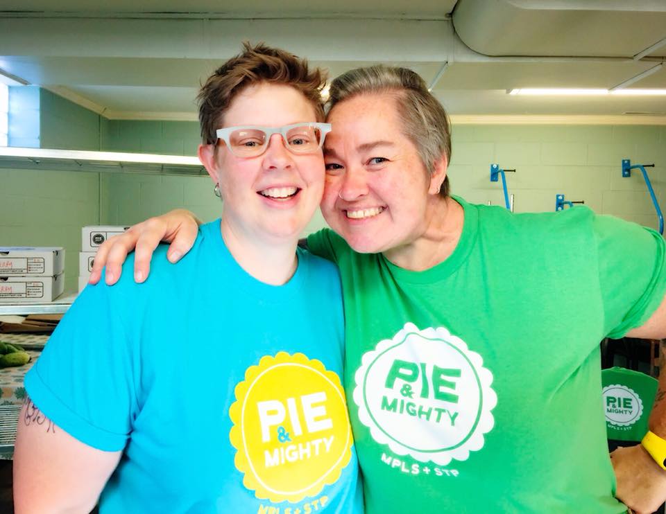Two women, one in glasses and a blue t-shirt another in a green t-shirt smile and embrace.