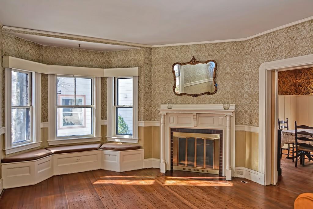 An empty parlor with a fireplace, a mirror above the fireplace, and a bay window.