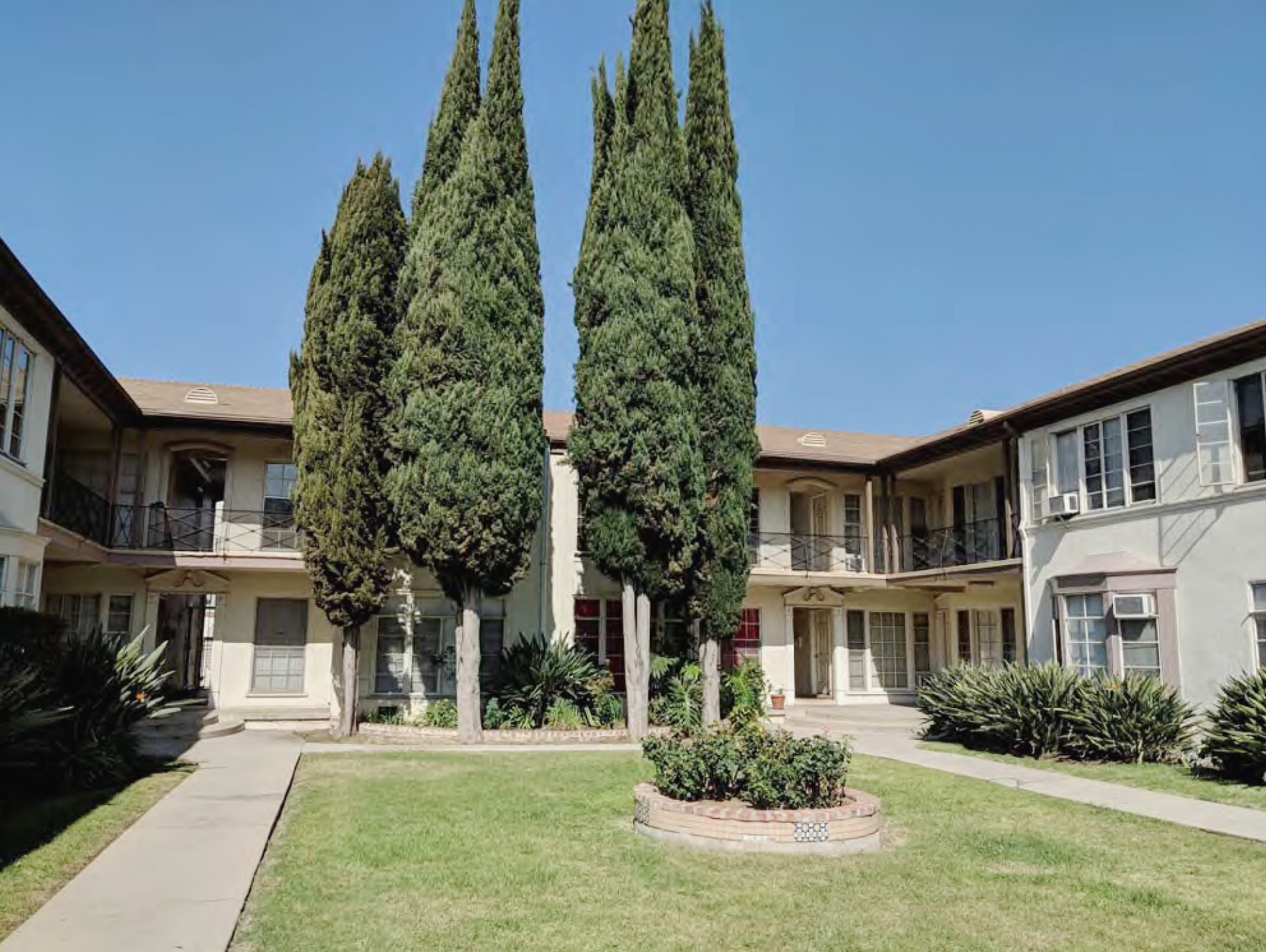 A photo of a green lawn surrounded on three sides by an apartment building. Tall cypress trees grow in a cluster at the far end of the lawn. 