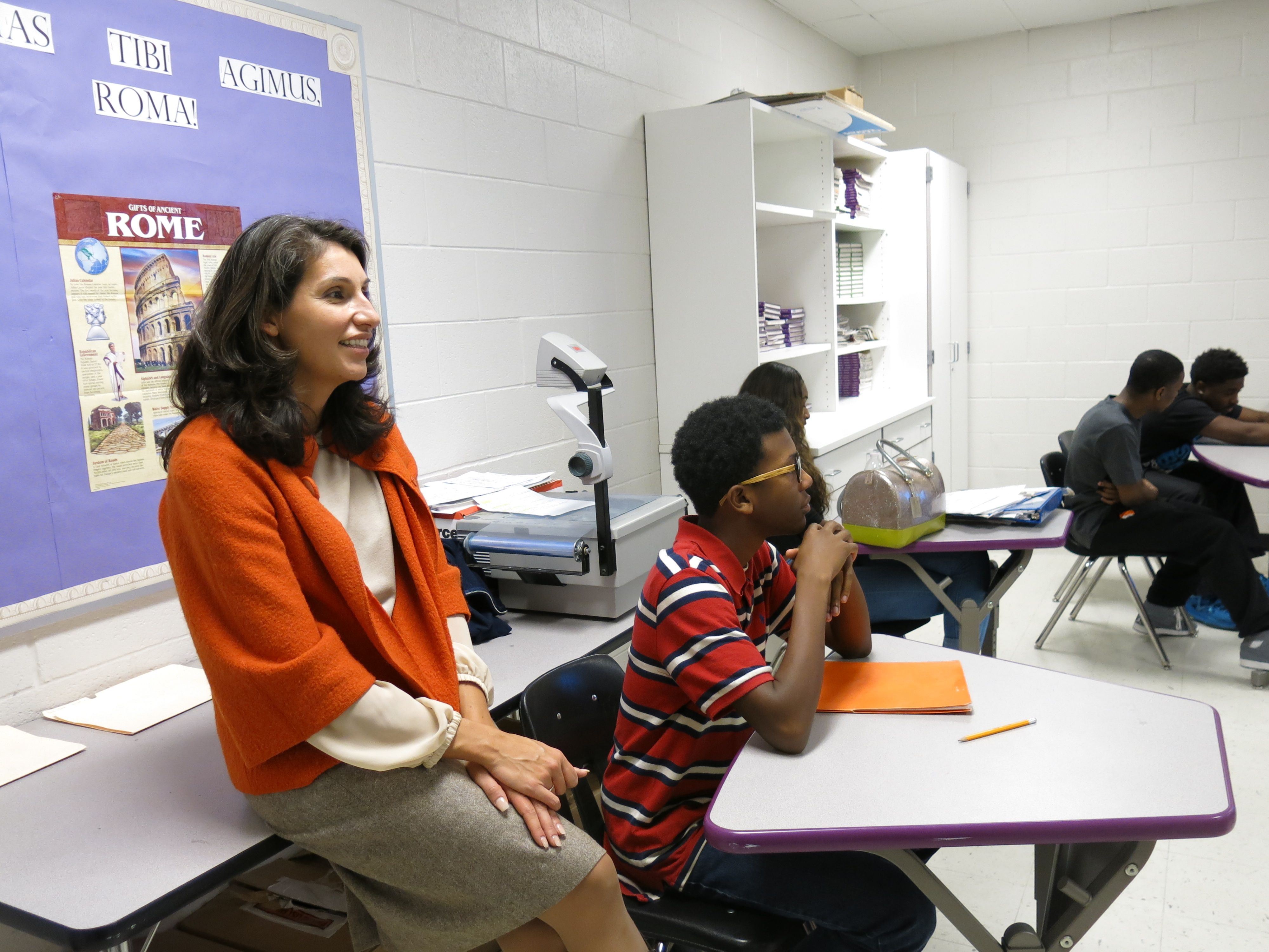 Heidi Ramirez, in an orange sweater and slacks, leans against a table at the back of a high school classroom while students sit at desks, on a visit in 2014 to Southwind High School in Memphis.