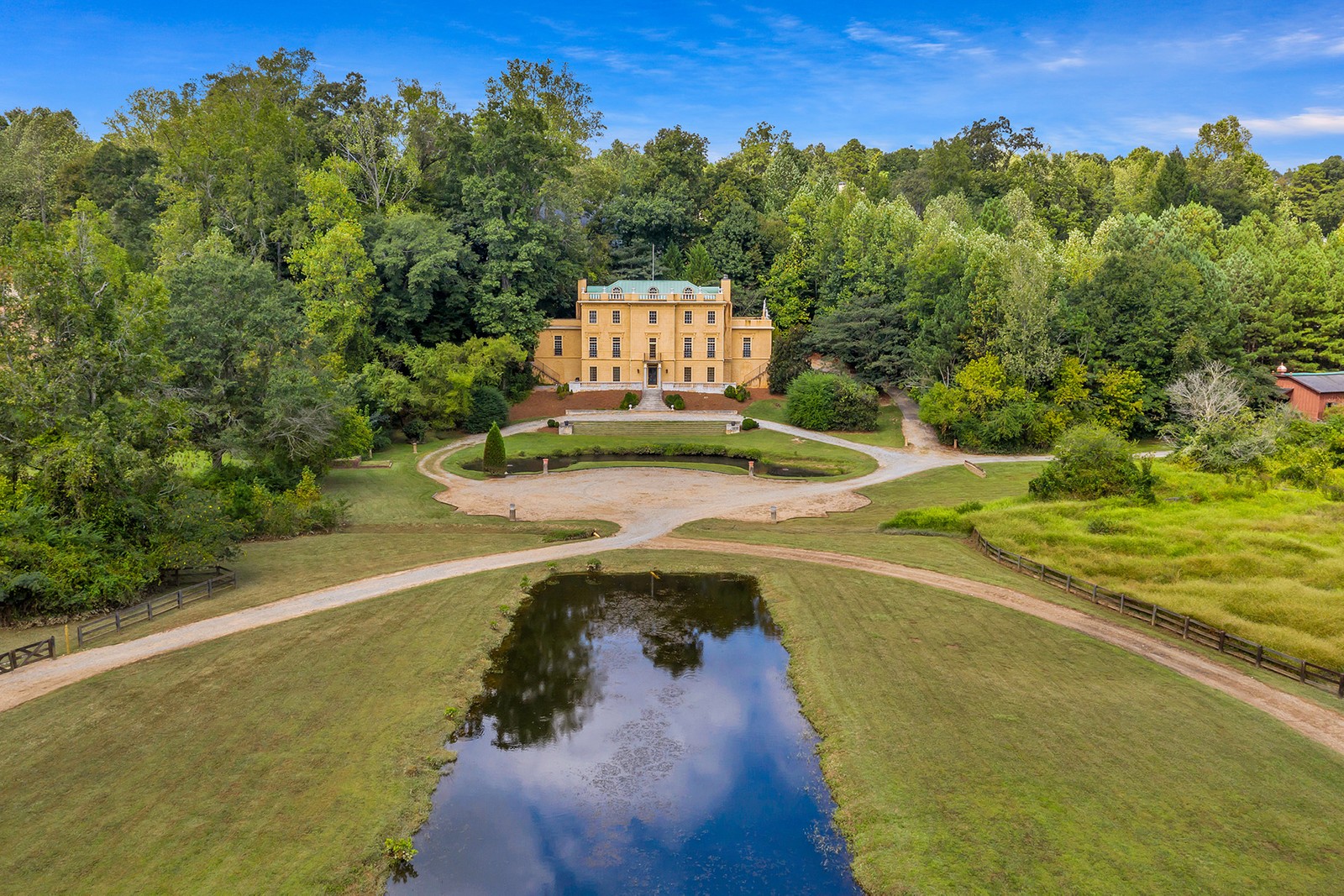 A yellow European-style mansion set atop a hill overlooking a pond with green grass all around. 