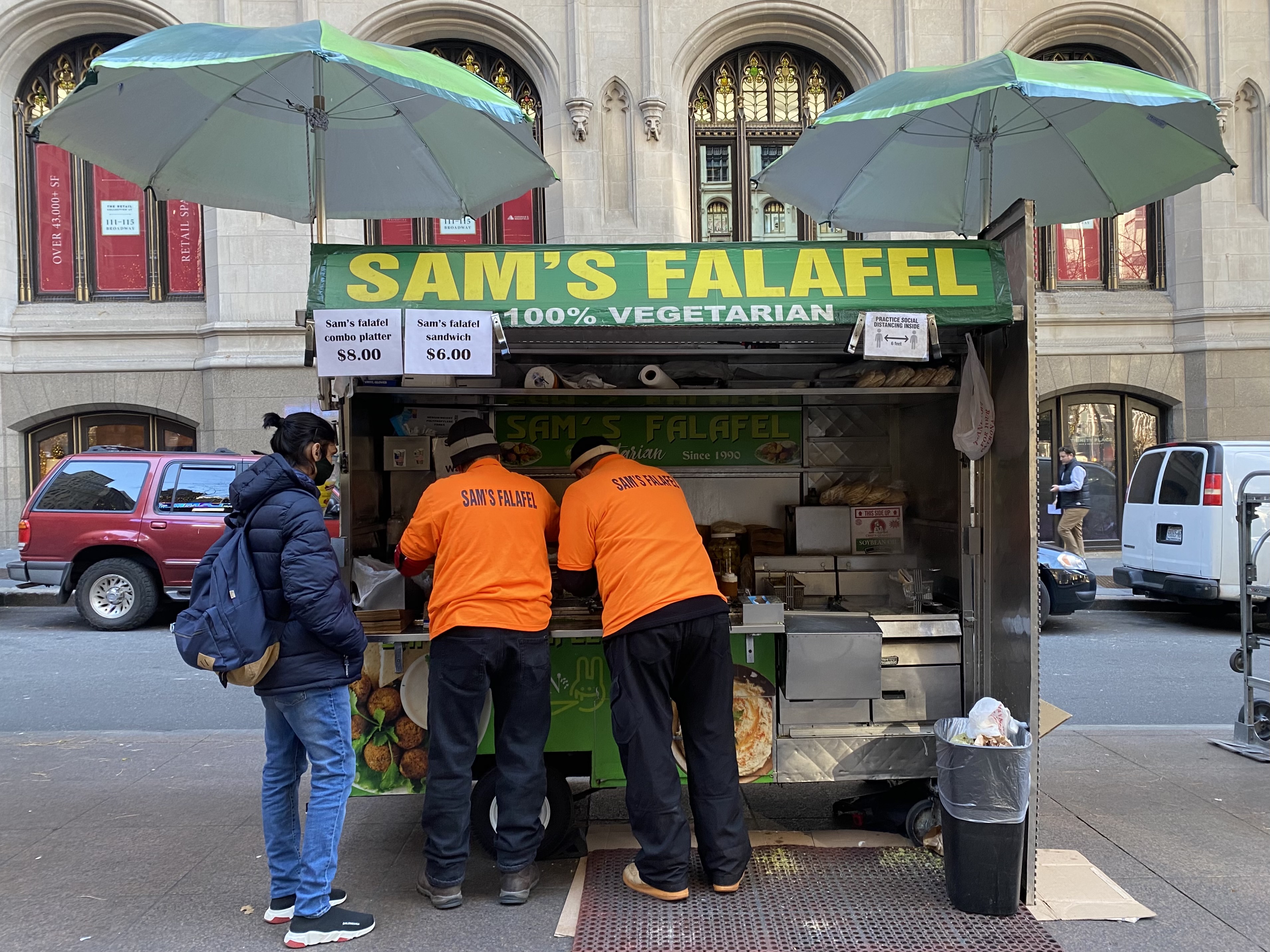 Two people wearing orange shirts with the words “Sam’s Falafel” in black lettering stand at an outdoor food cart.