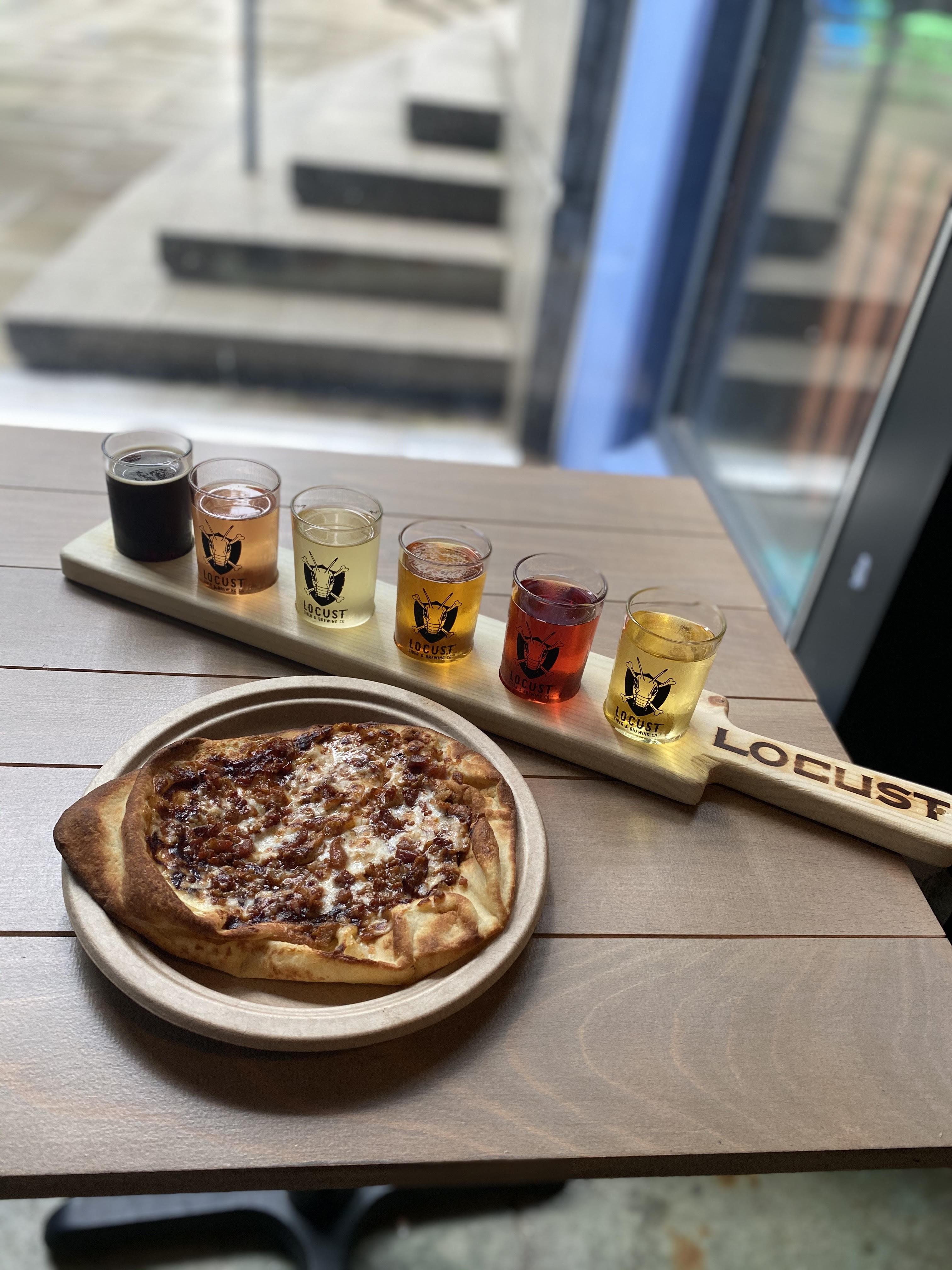 A wooden paddle with six cider tasters sits on a wooden table in the window at Locust’s Vancouver taproom, sitting next to a cheesy flatbread pizza on a base of lightly-charred naan
