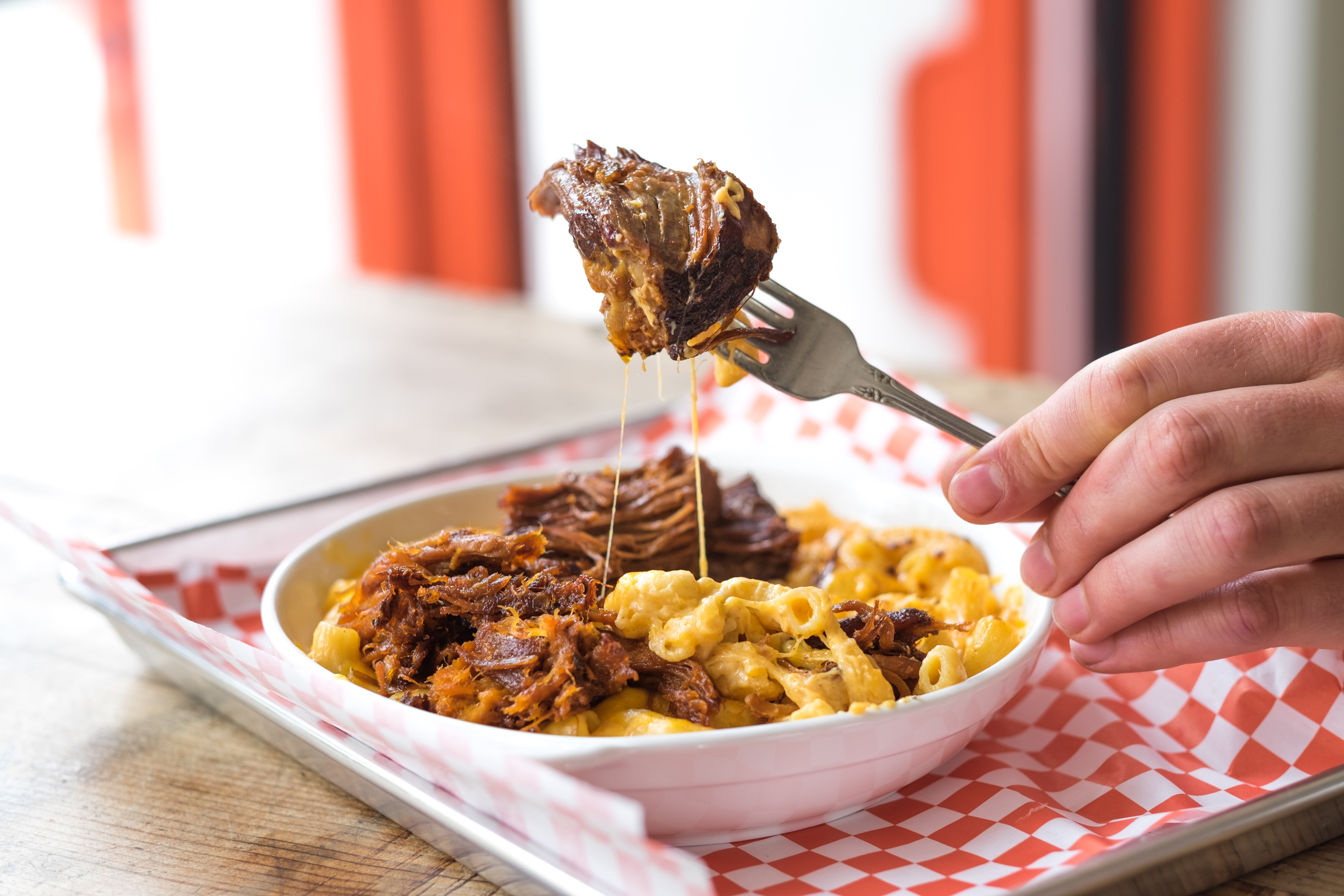 A fork holds up a hulking piece of brisket above a bowl of cheesy noodles and meat, sitting on a red-and-white-checkered paper on a metal tray