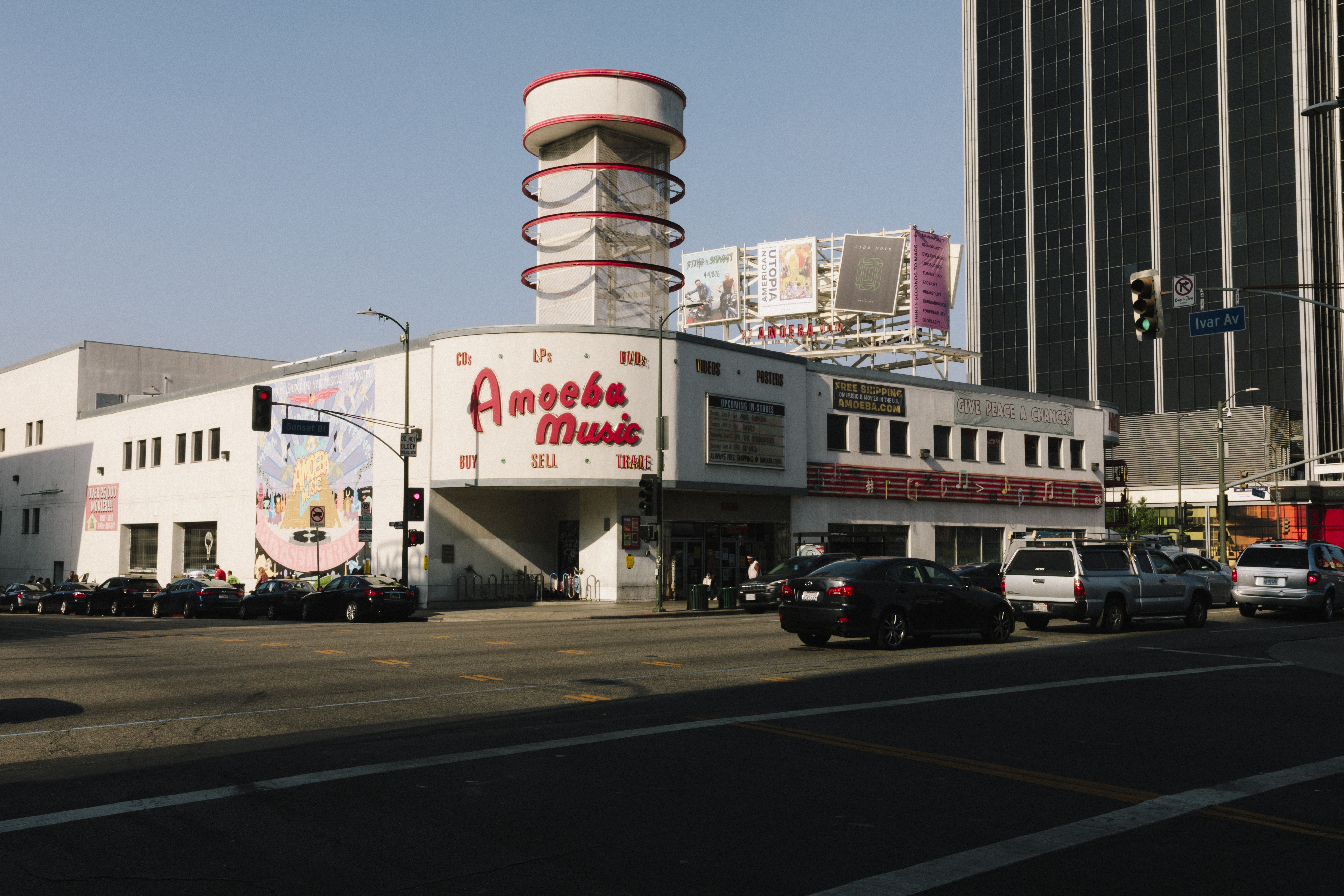 A photo of a low-rise, white, stucco building with exterior murals and neon signs, including one that says “Amoeba Records” in red letters on the front.