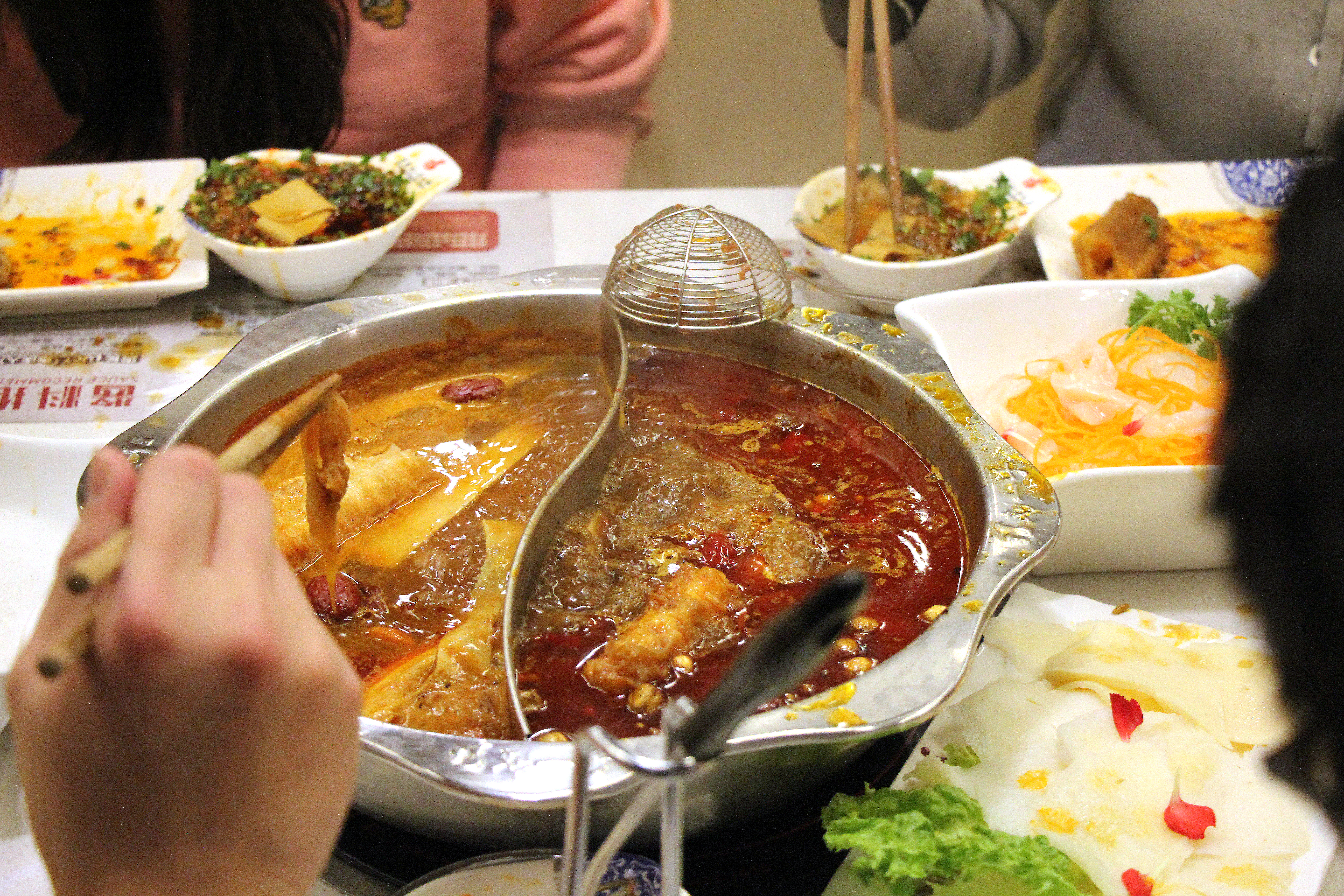 A hand lifts food from a silver hot pot bowl with a divider in the middle.